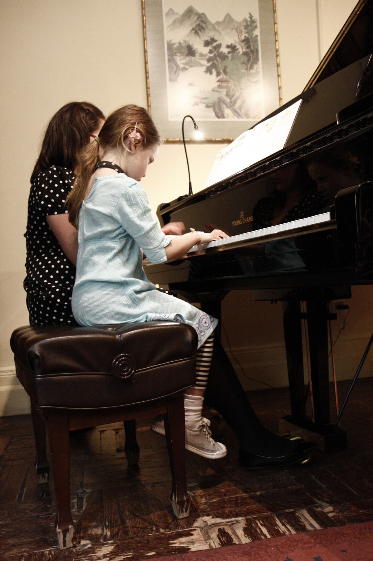 Two children, one in a light blue outfit, sit at a piano, with a woman nearby, in a room with artwork and a rug.