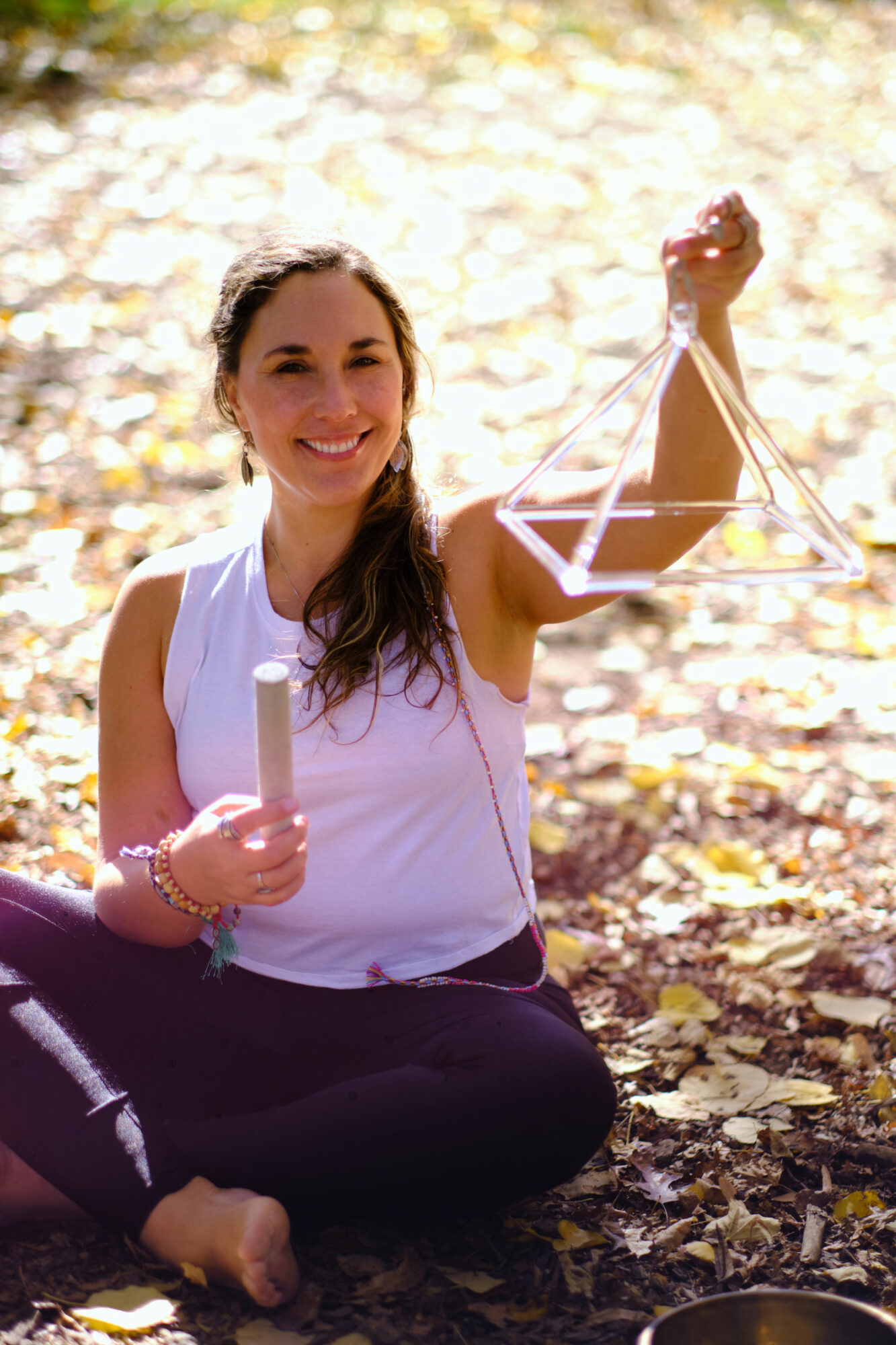 Young woman sitting outdoors on fallen leaves, smiling, holding a small scale in one hand and a tube in the other.