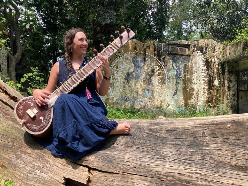 Woman sitting on a large log outdoors, holding a guitar, with trees and a stone wall in the background.