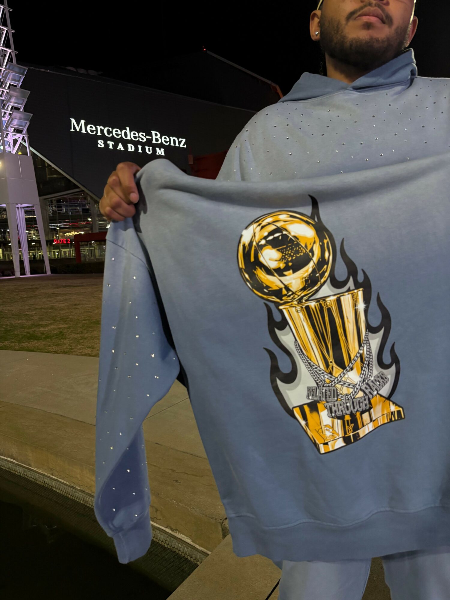 Person wearing a hoodie with a large graphic of a trophy and flames, standing indoors near a Mercedes-Benz display.