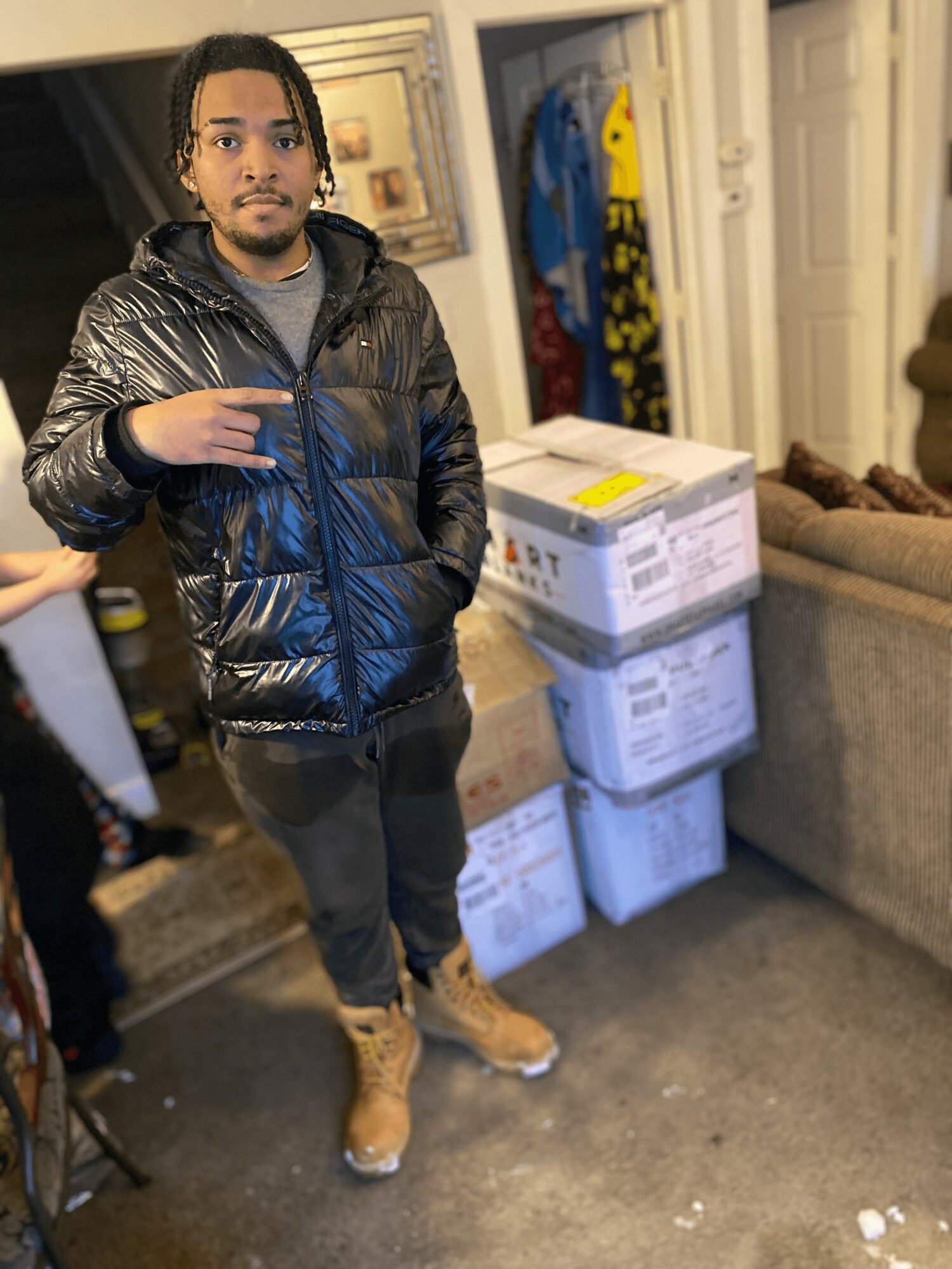 Young person with dark hair and a black puffer jacket standing indoors near boxes and a sofa.