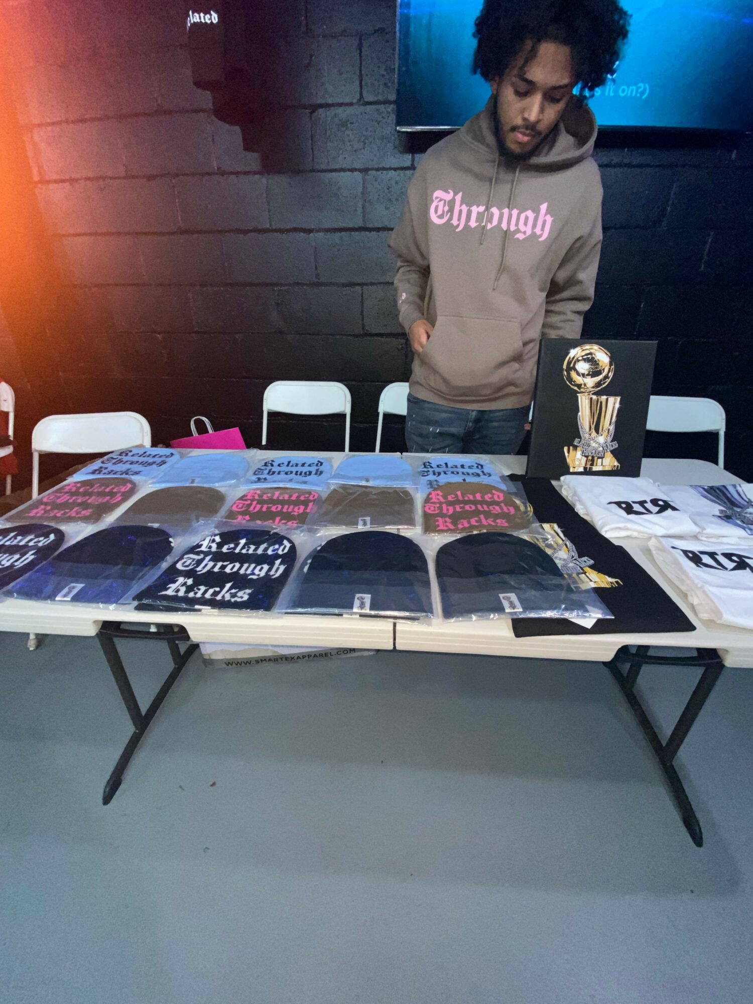 Person standing behind a table with various printed items, trophies, and a sign, in an indoor setting.