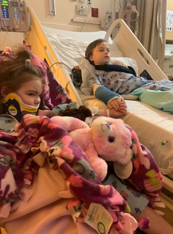 Young girl lying in hospital bed holding pink stuffed animal, surrounded by toys and blankets.