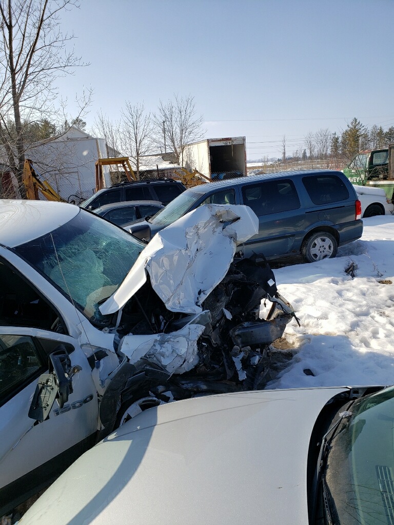 Car crash scene with damaged vehicle and snow-covered ground, multiple parked cars, and trees in the background.