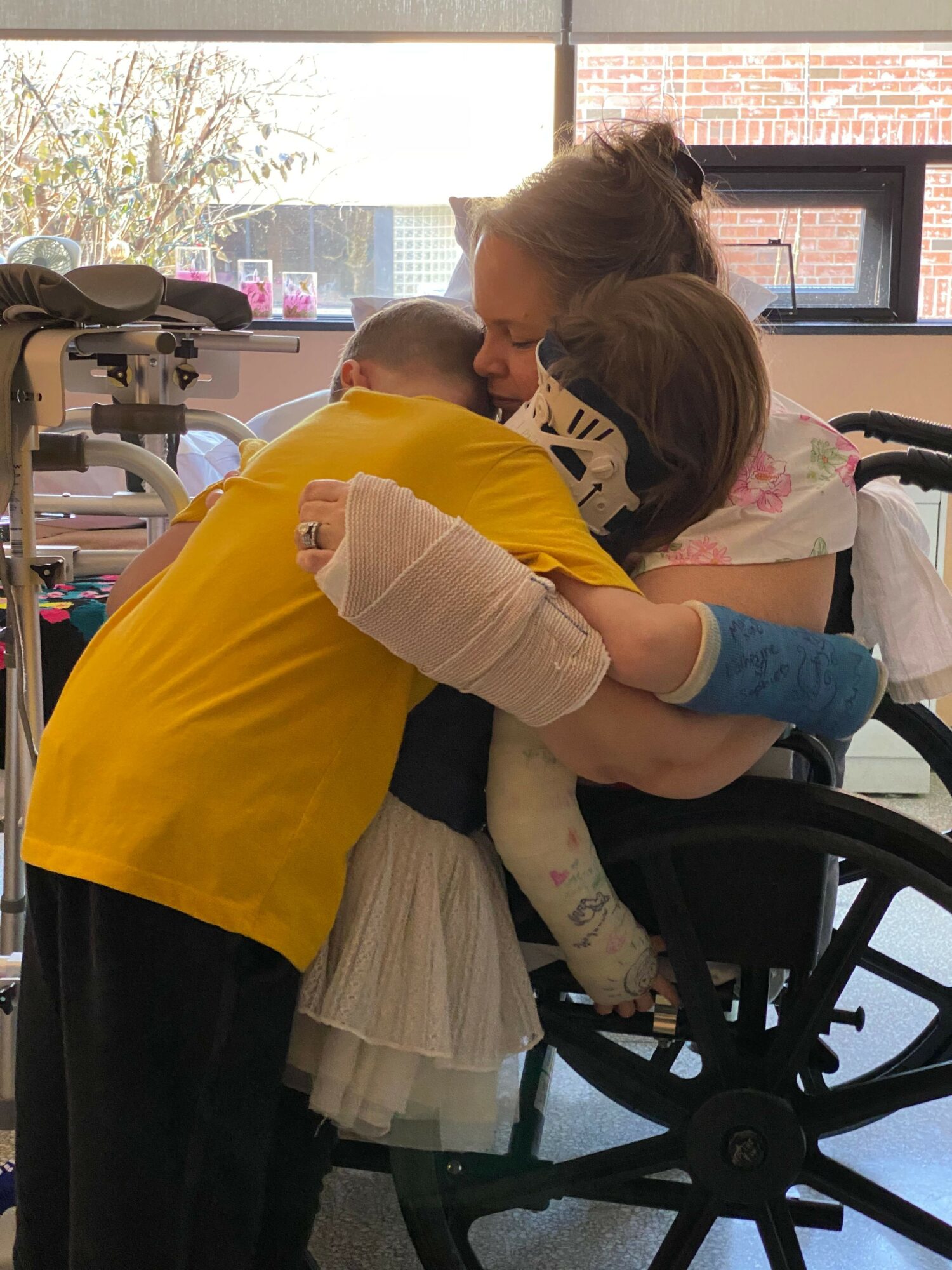 Child in a yellow shirt hugging a person in a yellow shirt, near a wheelchair inside a room with large windows.