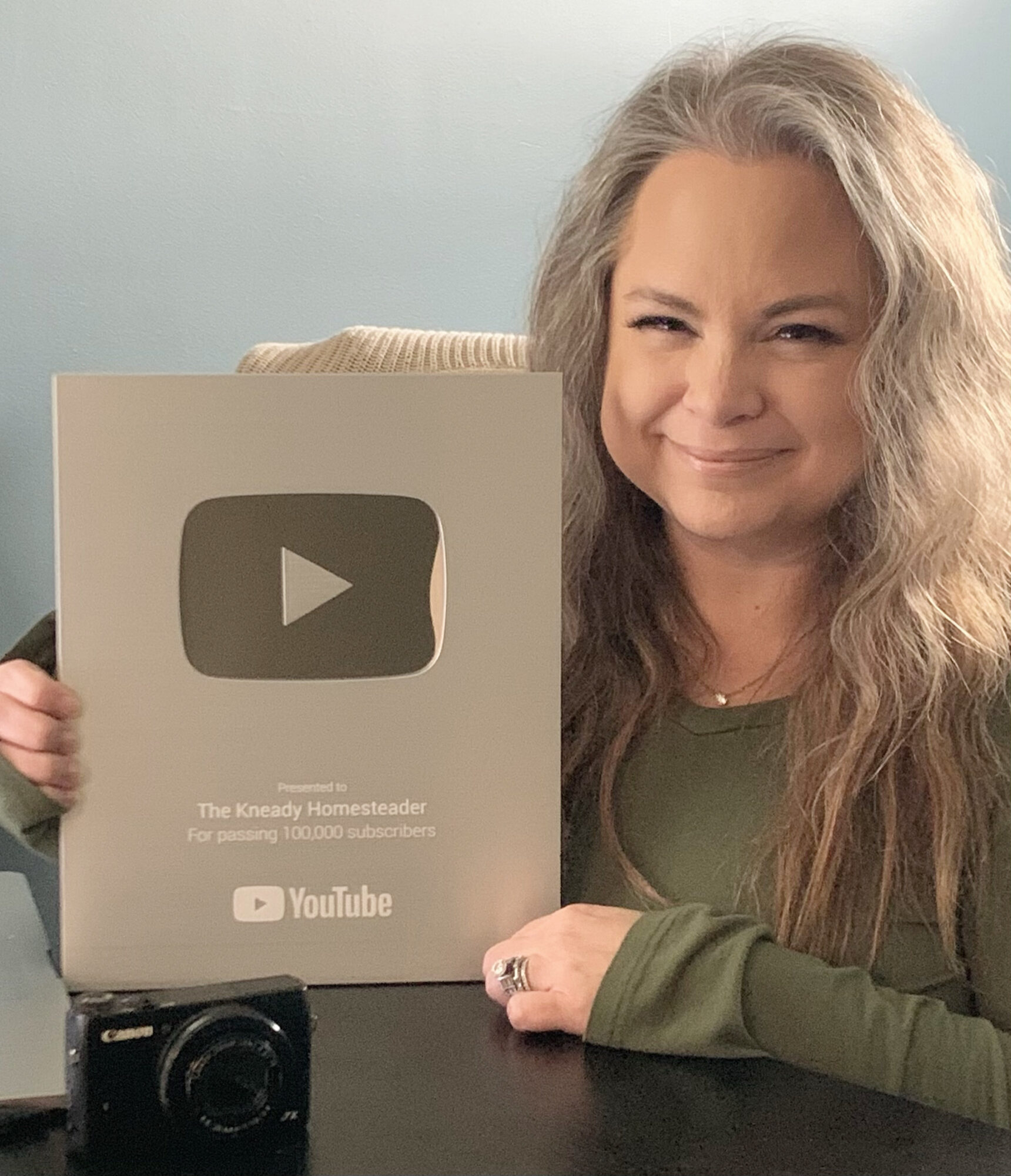 Woman with long gray hair holding a YouTube Silver Play Button award, smiling, in a room with light-colored wall.
