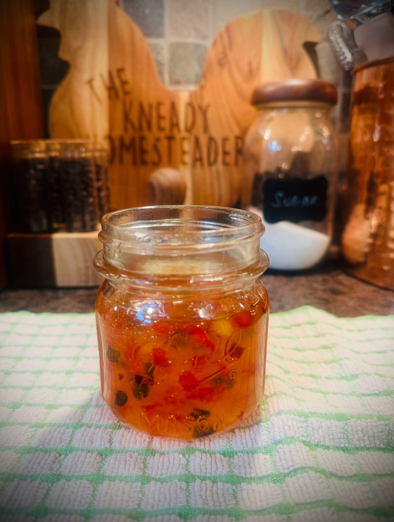 Small glass jar with red and black berries inside, placed on a green and white striped cloth, with kitchen items in background.