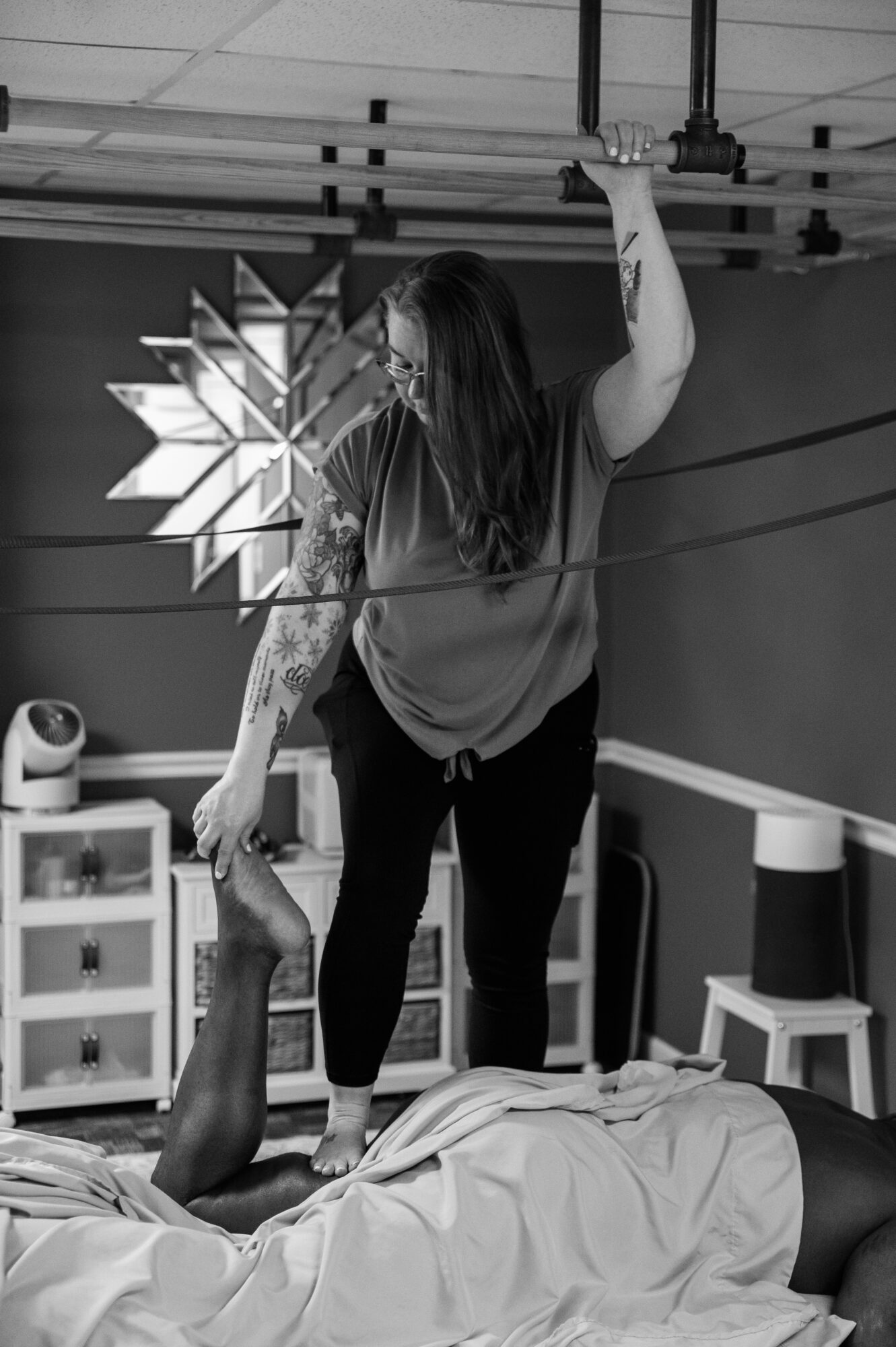 A woman standing on a bed, holding a pole, with a person lying down beneath her in a room with furniture and a decorative wall piece.
