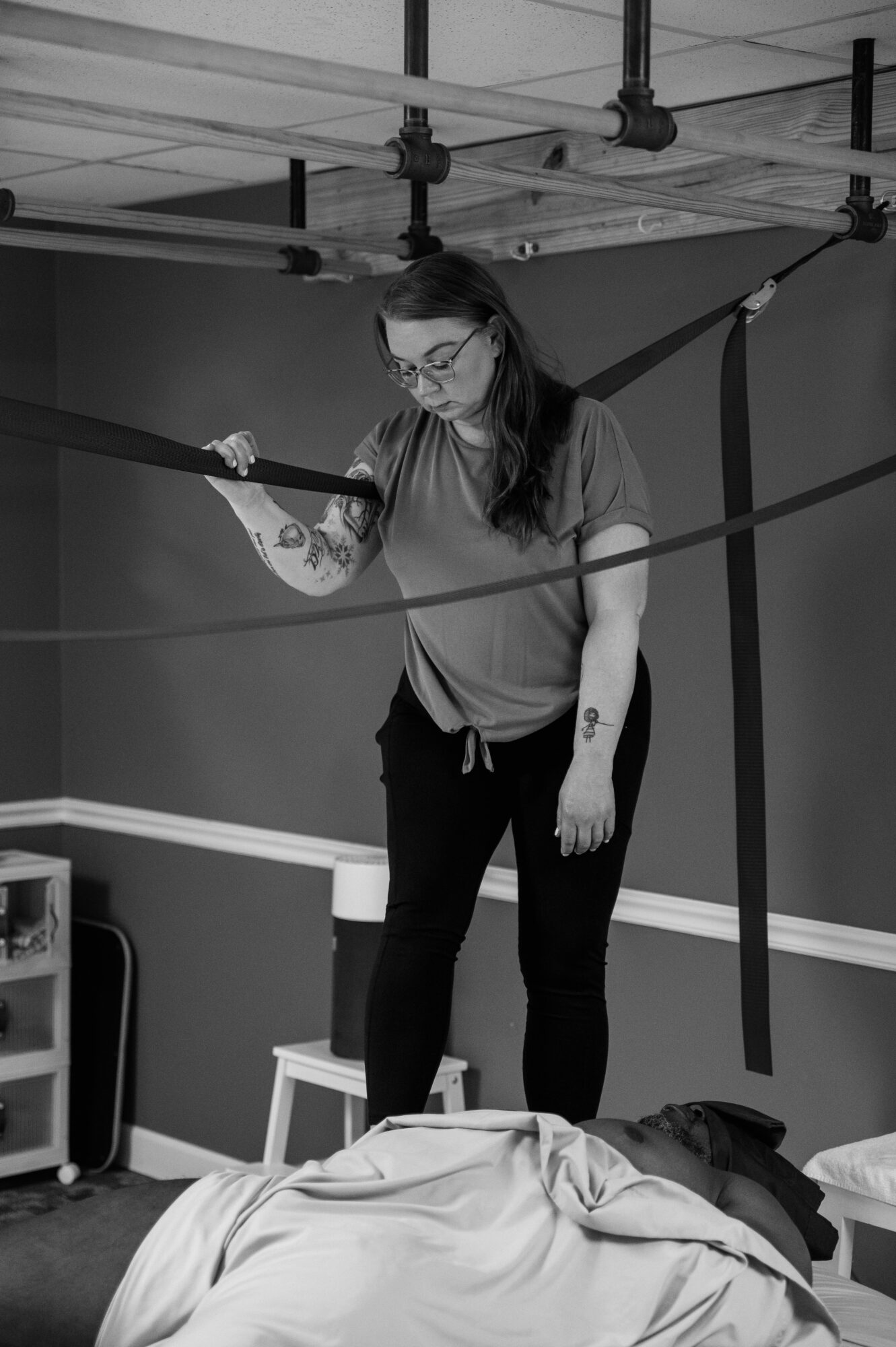 Woman with glasses standing on a bed, holding a strap, in a room with ceiling pipes and a speaker, in black and white.
