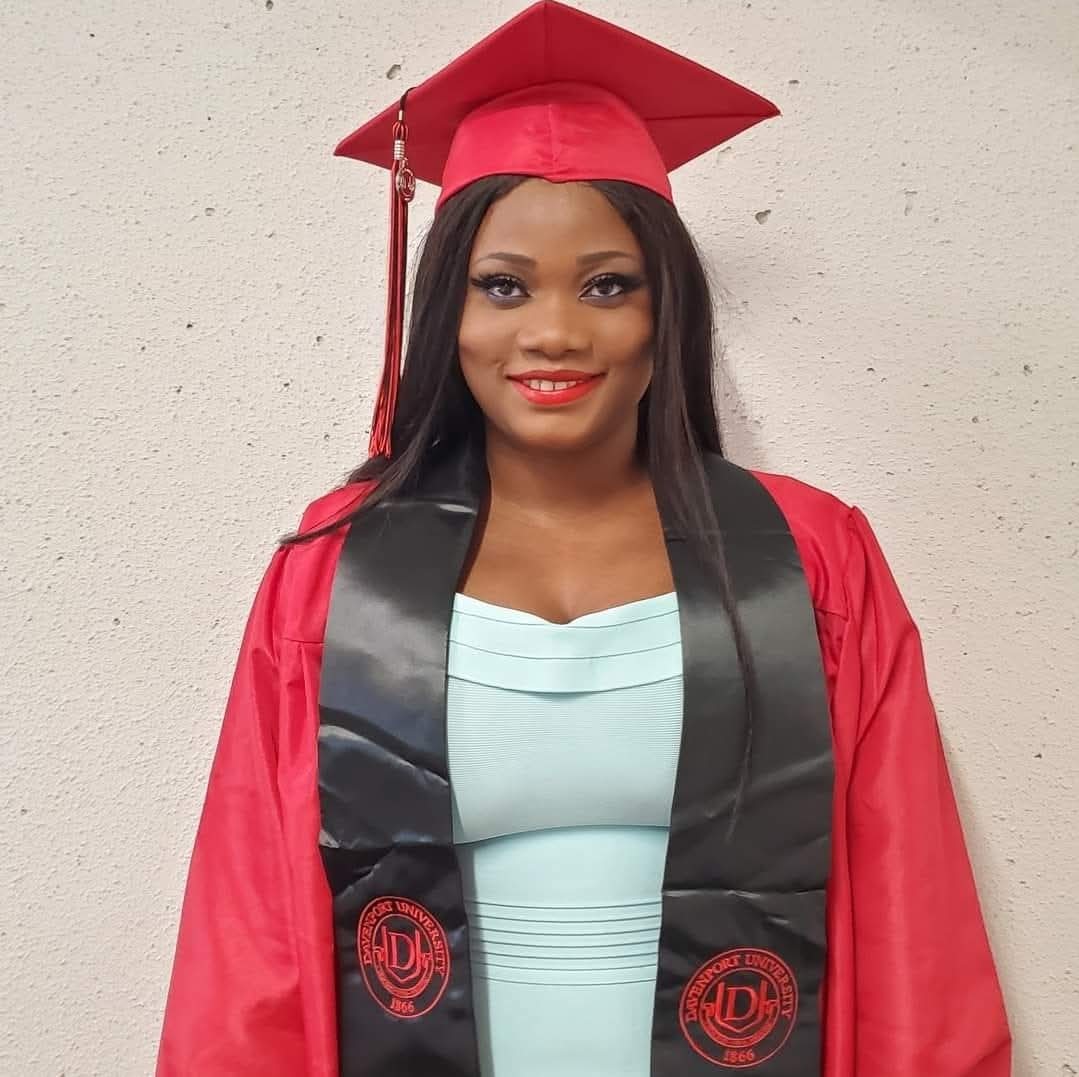 Young woman in graduation cap and gown smiling, standing against a plain wall.