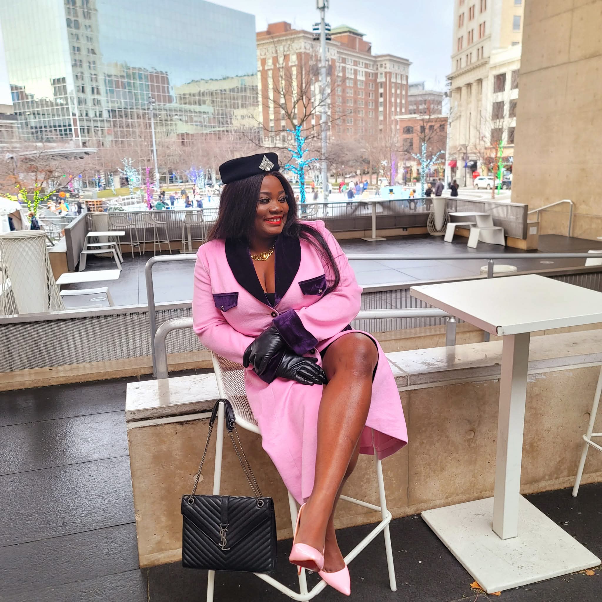 Woman in pink coat and black gloves sitting outdoors with city buildings and trees in background.