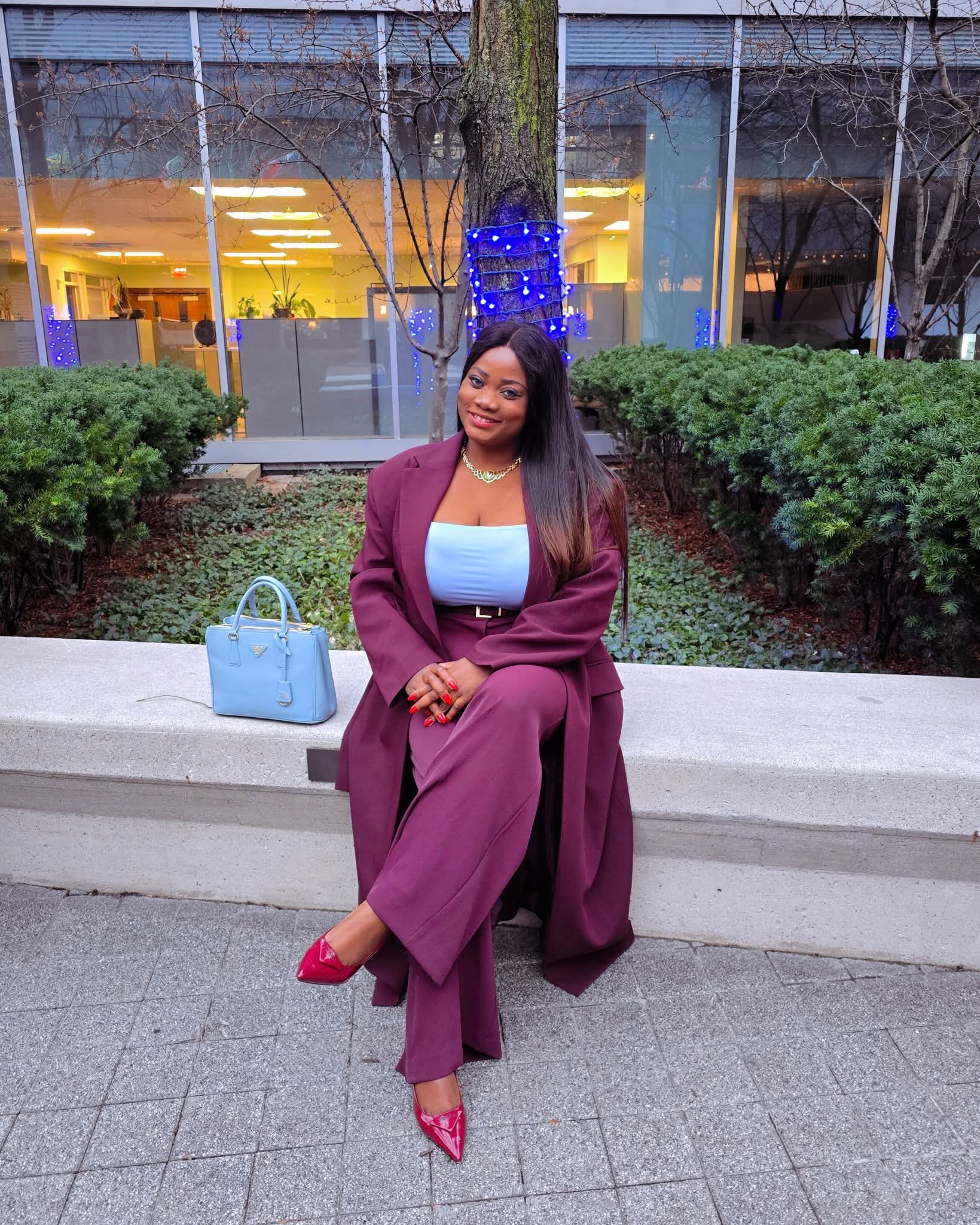 Woman sitting on a concrete ledge outdoors, wearing a purple suit, pink shoes, and smiling, with a blue bag nearby.