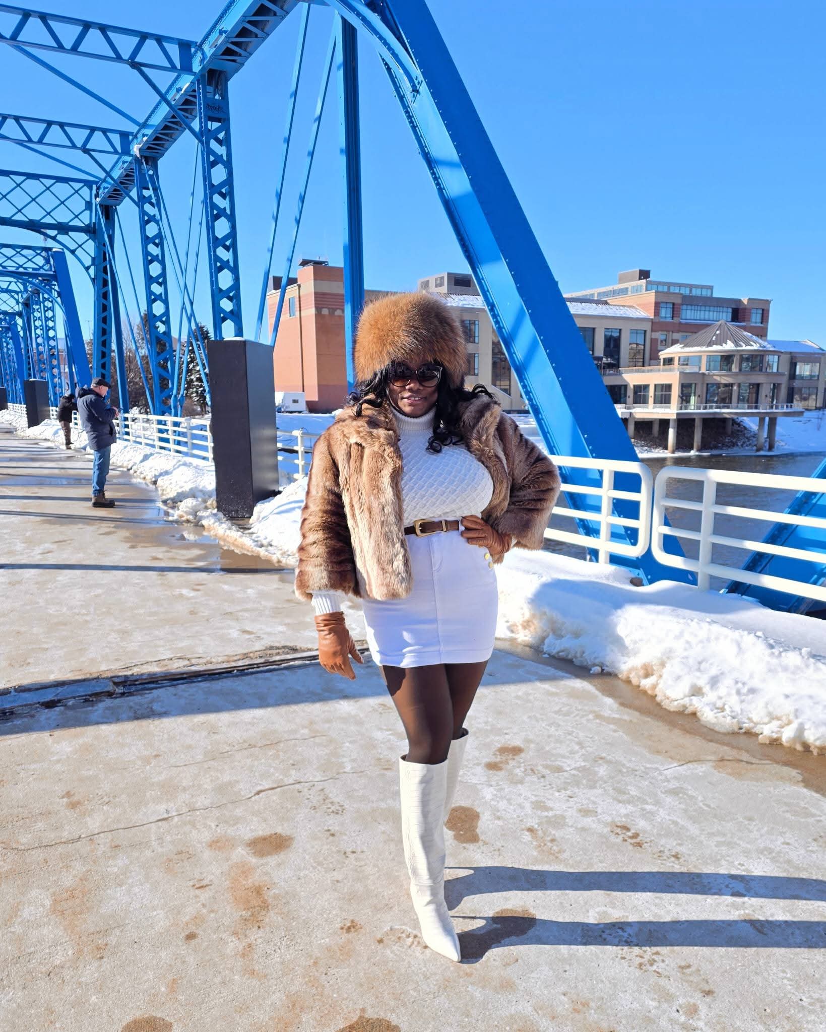 Woman in fur coat and white outfit walking on a bridge with blue structure, snow on sides, clear sky.