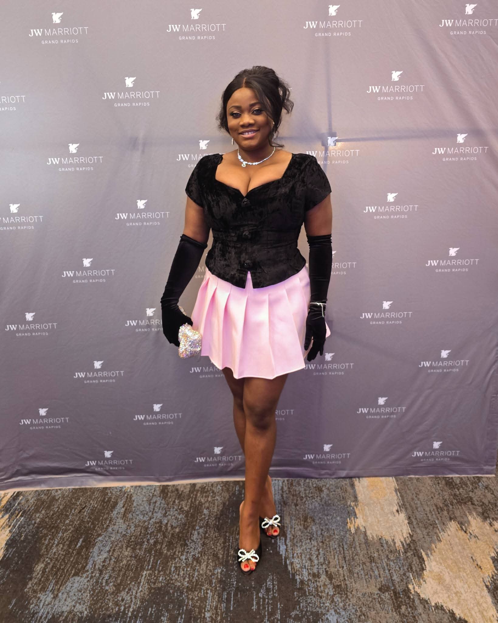 Woman in black top and pink skirt standing on carpeted floor against a gray backdrop, smiling, holding a small purse.