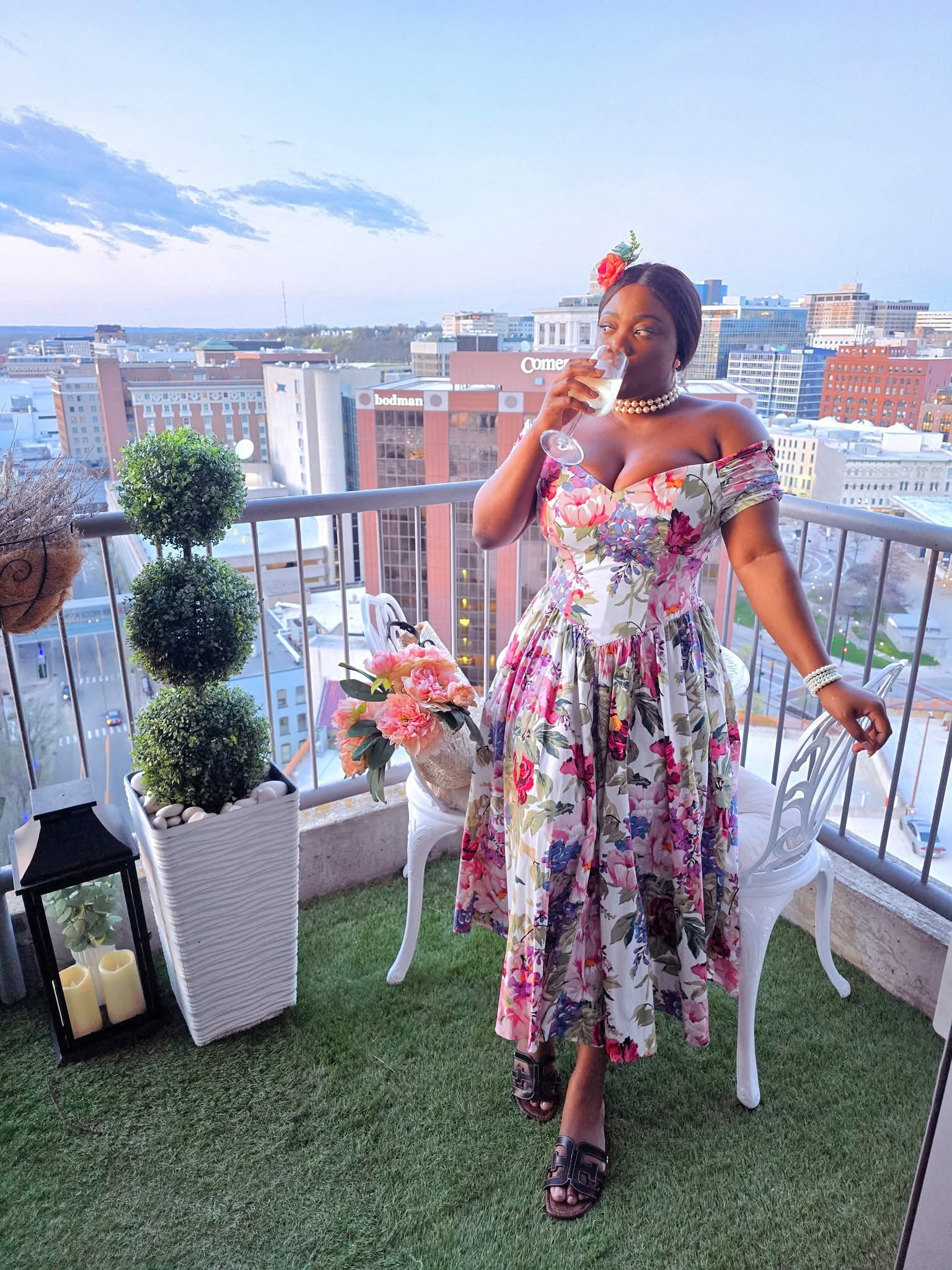 Woman in floral dress drinking from a glass on a balcony with city view, plants, and outdoor furniture.