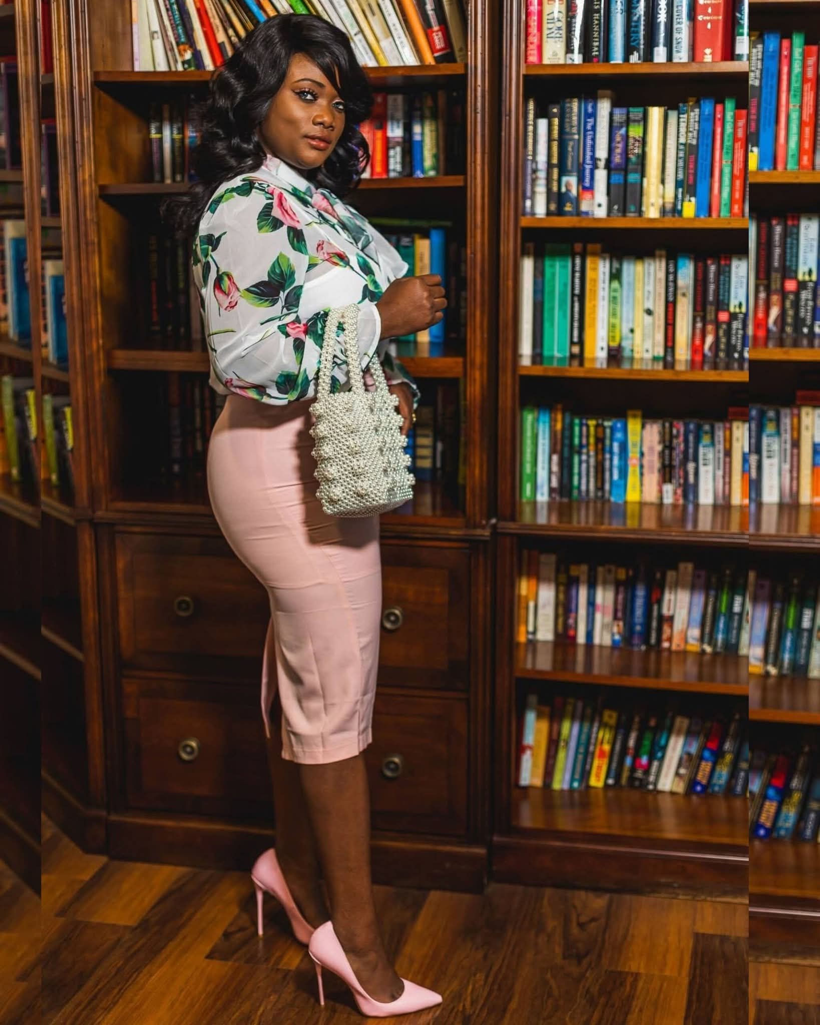 Woman in floral blouse and pink skirt standing in front of bookshelves holding a bag, in a library setting.