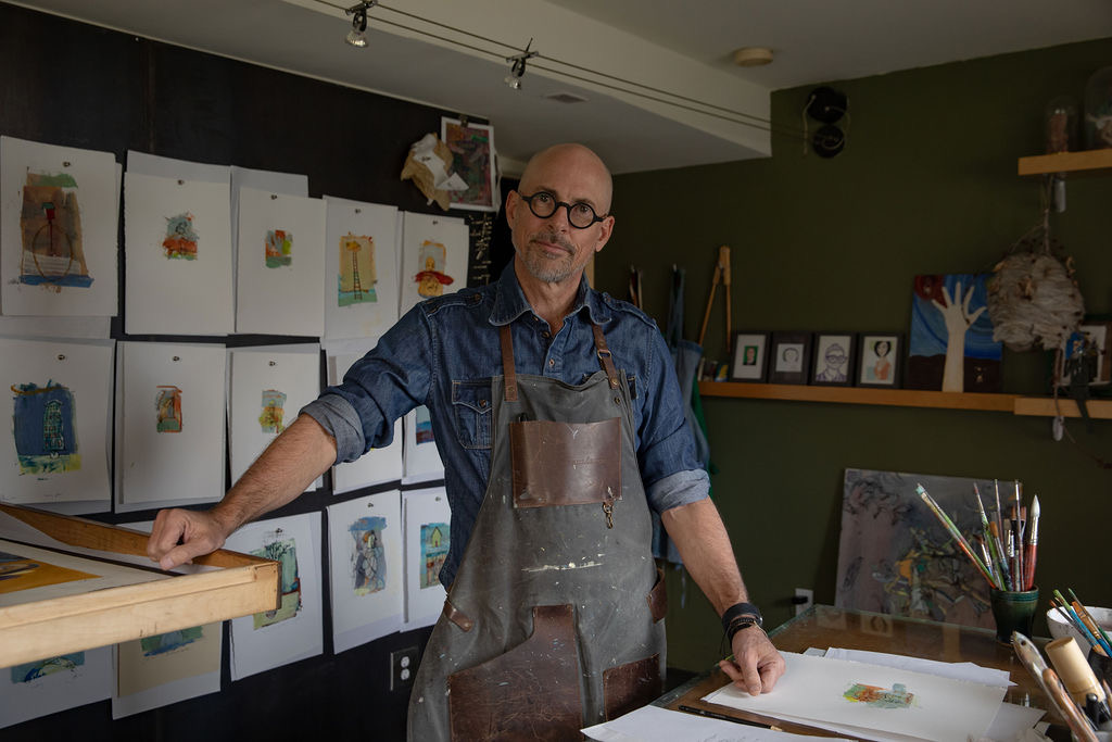 Man with glasses and apron standing in art studio with paintings and supplies, smiling at camera.