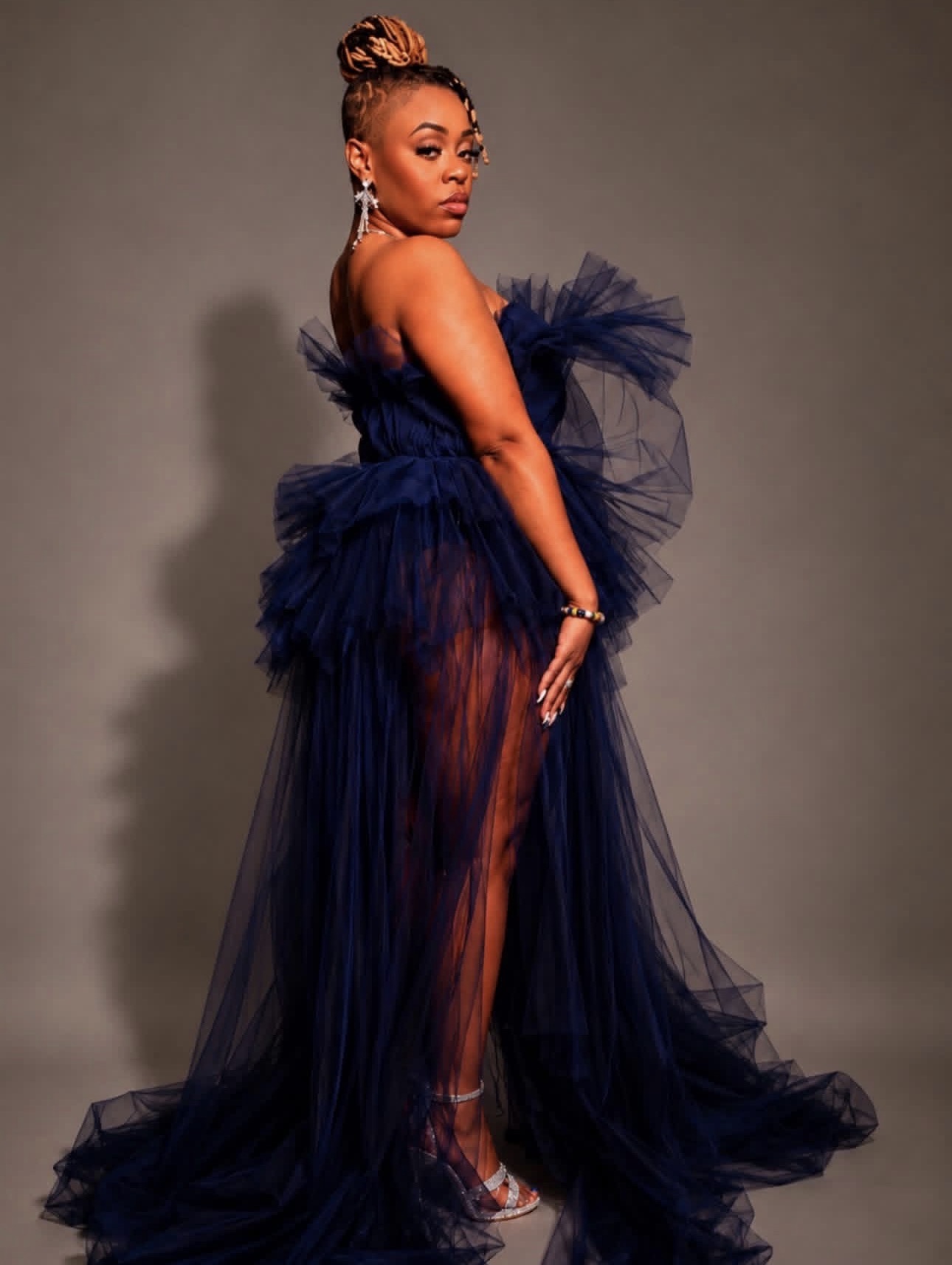 Woman in a dark blue tulle gown with high slit, standing sideways, looking over shoulder, with styled hair and jewelry.