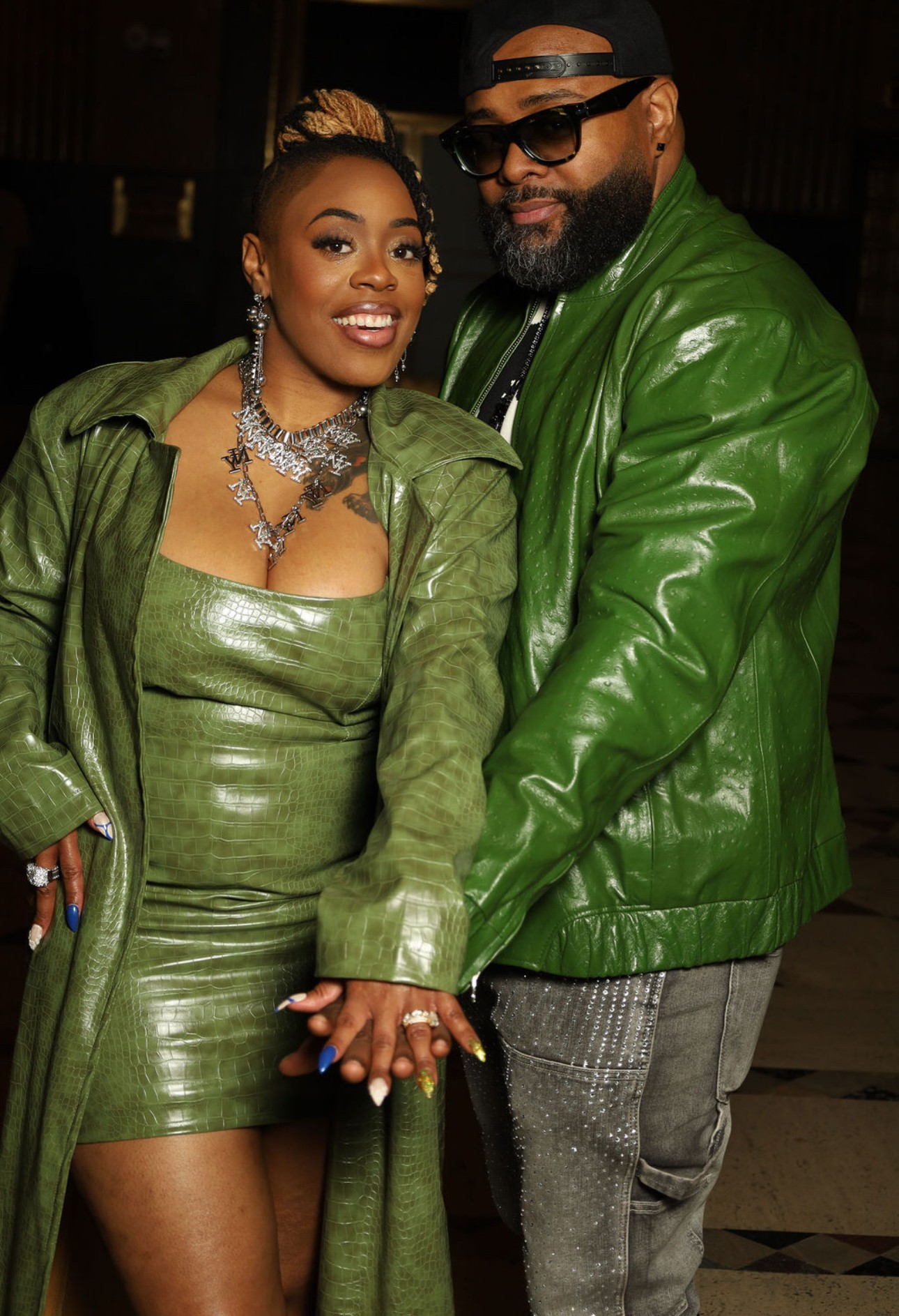 A woman and man pose together, both wearing green leather outfits, smiling, in a dimly lit setting.
