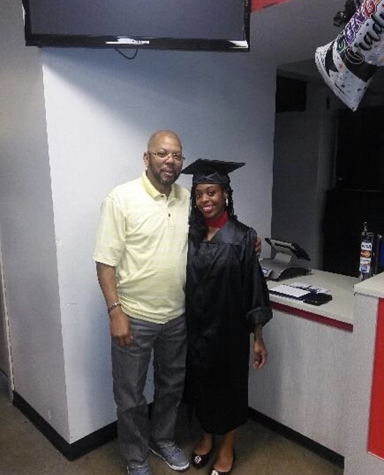 A man and woman in graduation attire standing indoors, smiling, with a TV and counter in the background.