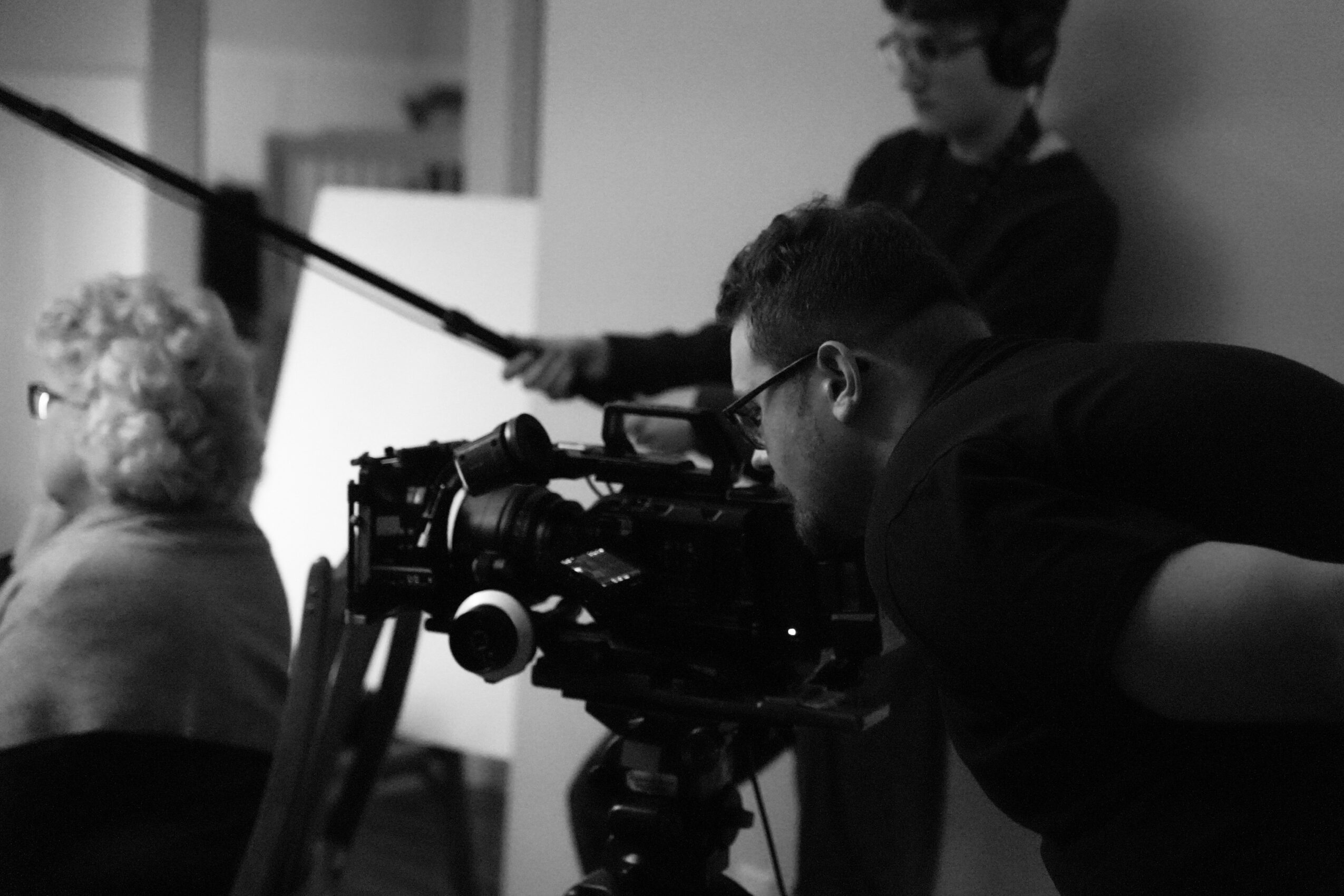 Two men operate a professional camera on a tripod, filming an elderly woman with curly hair in an indoor setting.