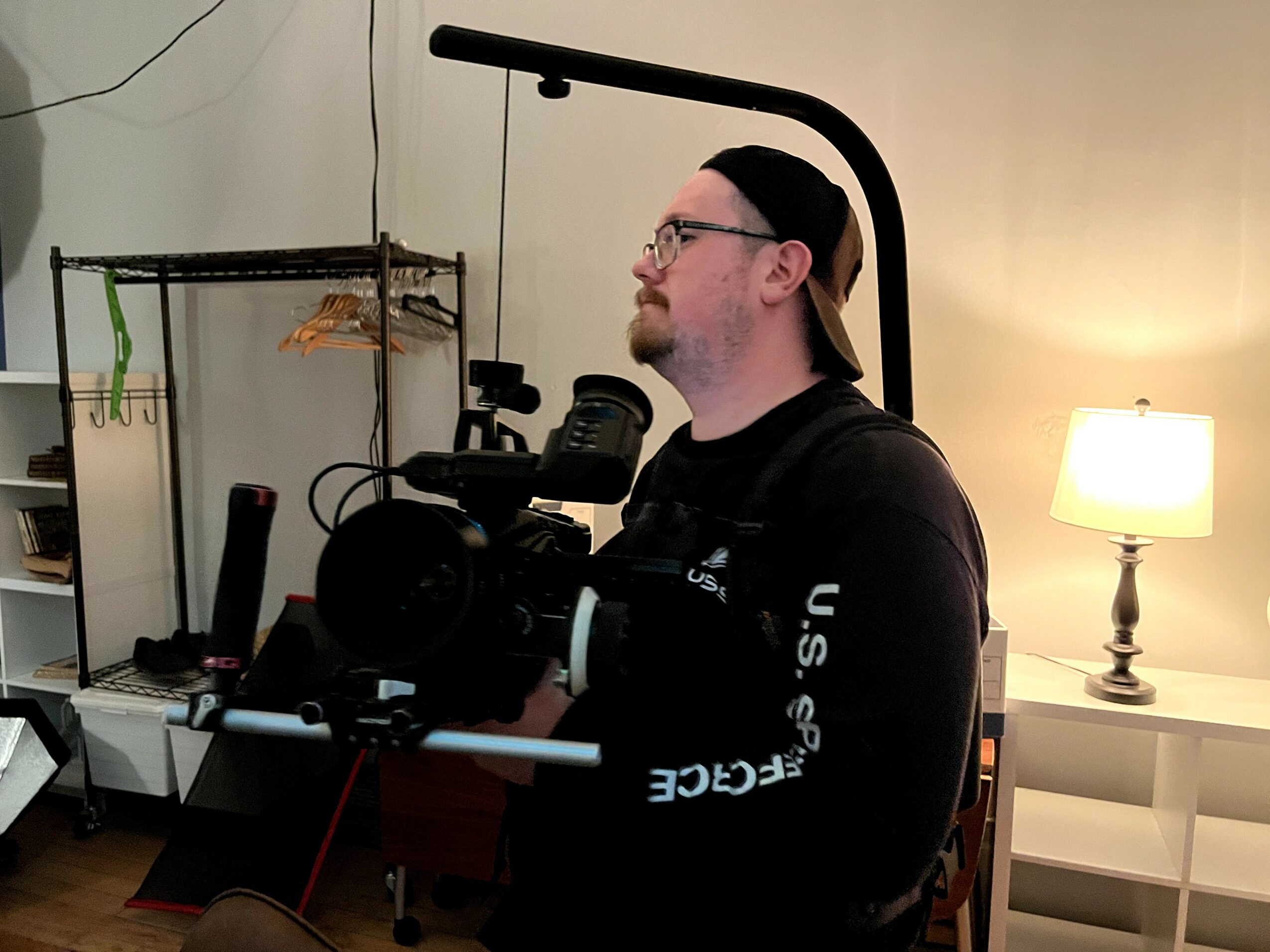 Person with glasses and a beard looking to the right, holding a camera in a room with a lamp and clothing rack