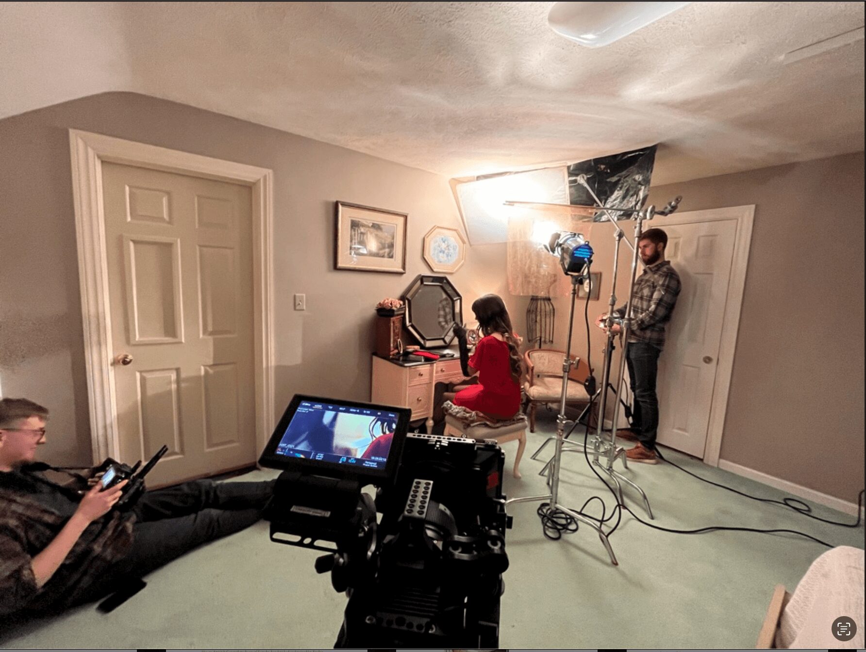 Person in red seated in front of camera, others with equipment in a room with closed door and wall art.