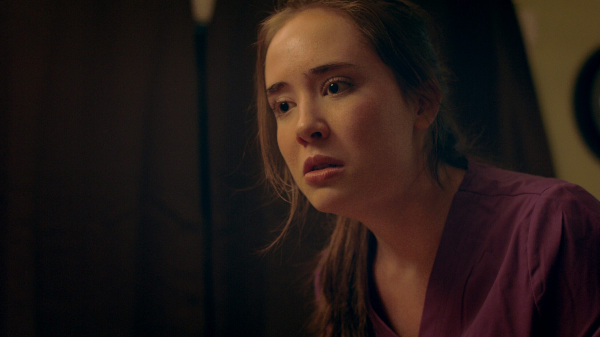 Young woman with long hair and a concerned expression, wearing a purple top, indoors with dark background.