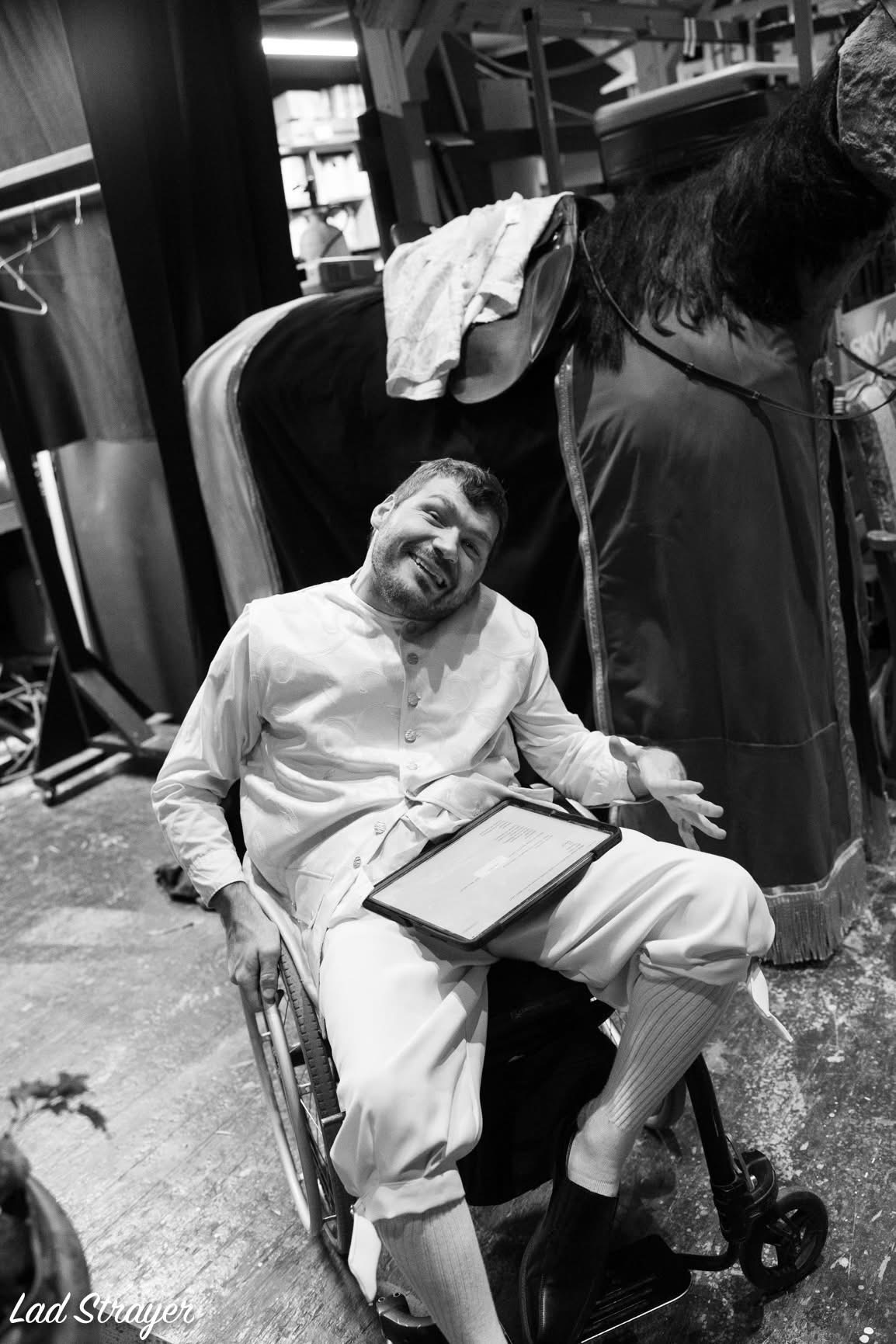 A man in a wheelchair smiling, holding a tablet, with a person leaning over behind him in a busy indoor setting.