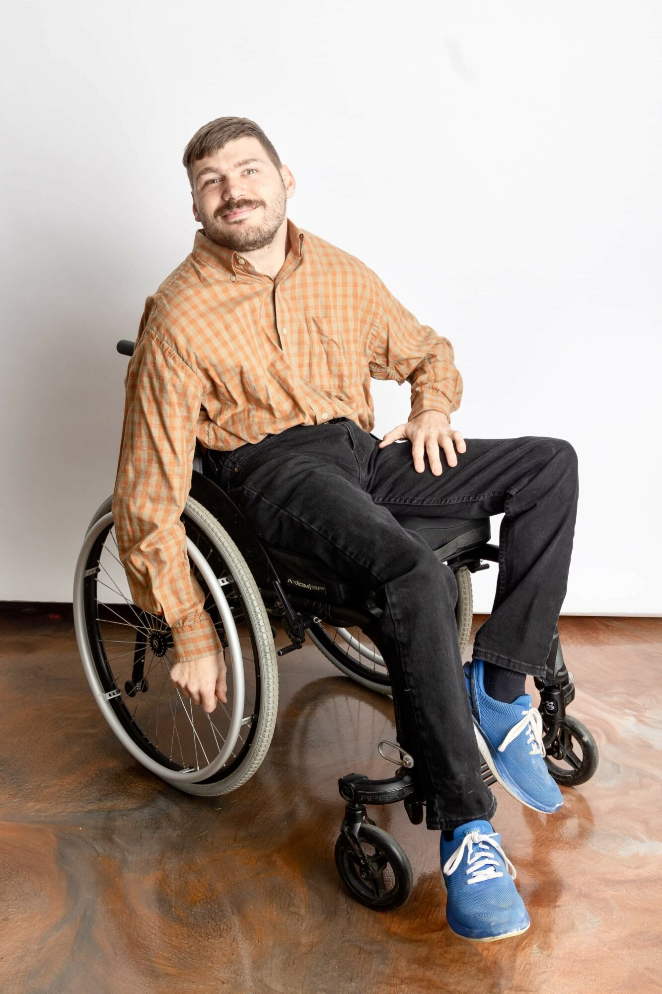 Man in wheelchair smiling, wearing orange checkered shirt, black pants, and blue sneakers, sitting on wooden floor against white wall.
