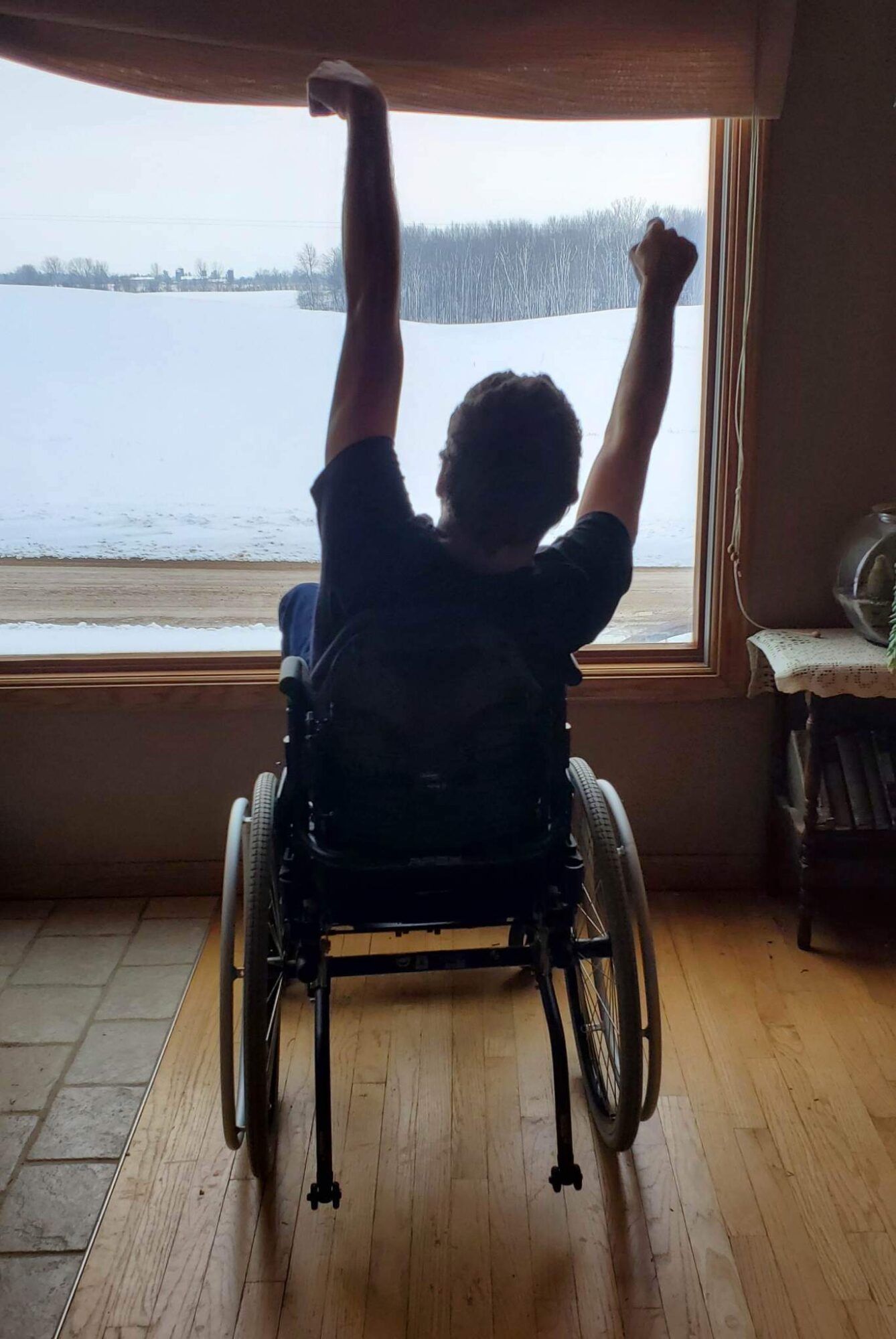 Person in a wheelchair with arms raised, facing a window overlooking a lake and trees, indoors with wooden floor.