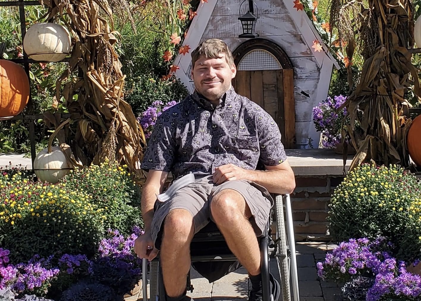 Young man in a wheelchair smiling outdoors with colorful flowers and a small house in the background.
