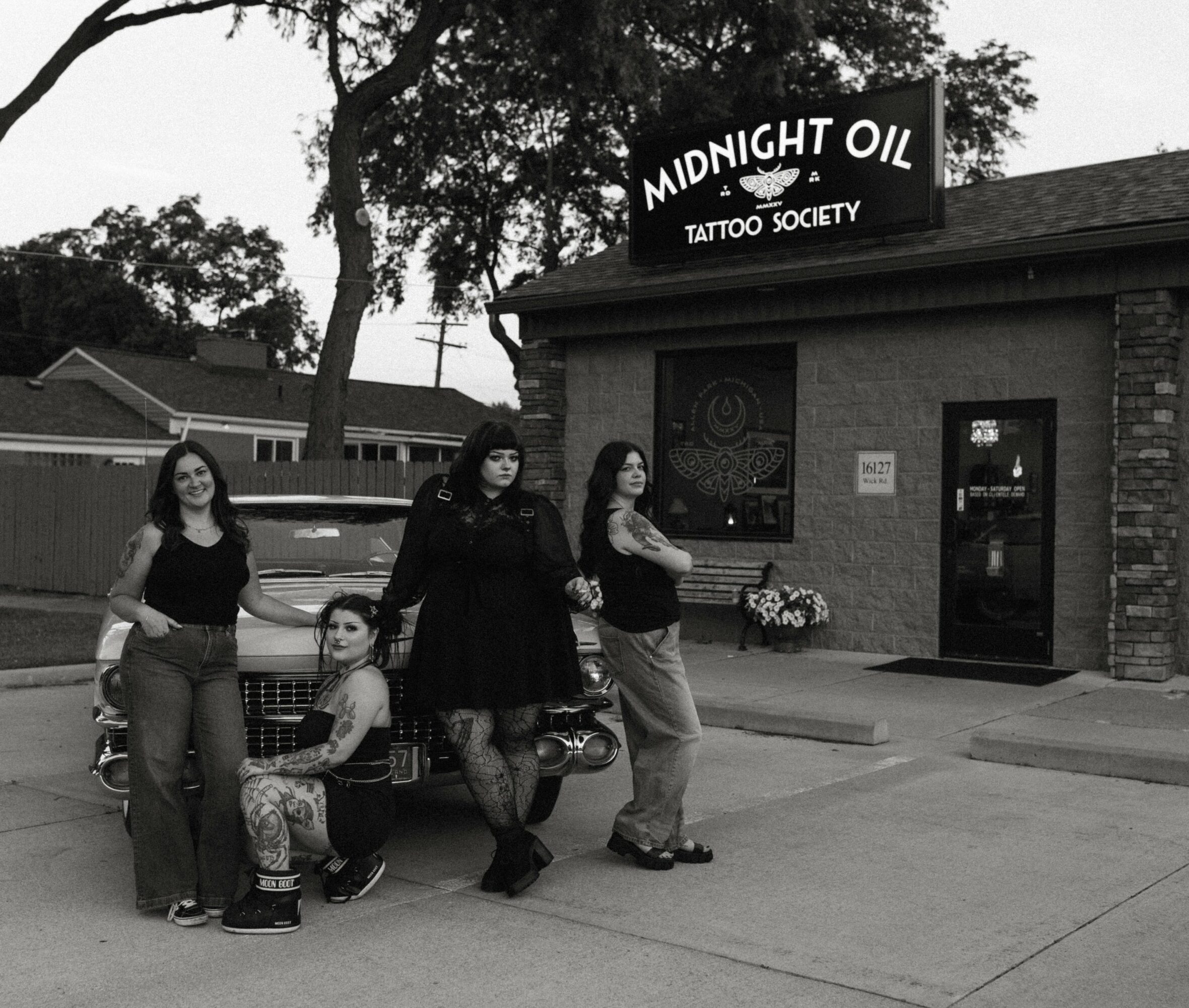 Four women and a girl stand outside a tattoo shop named Midnight Oil Tattoo Society, with trees and houses in background.