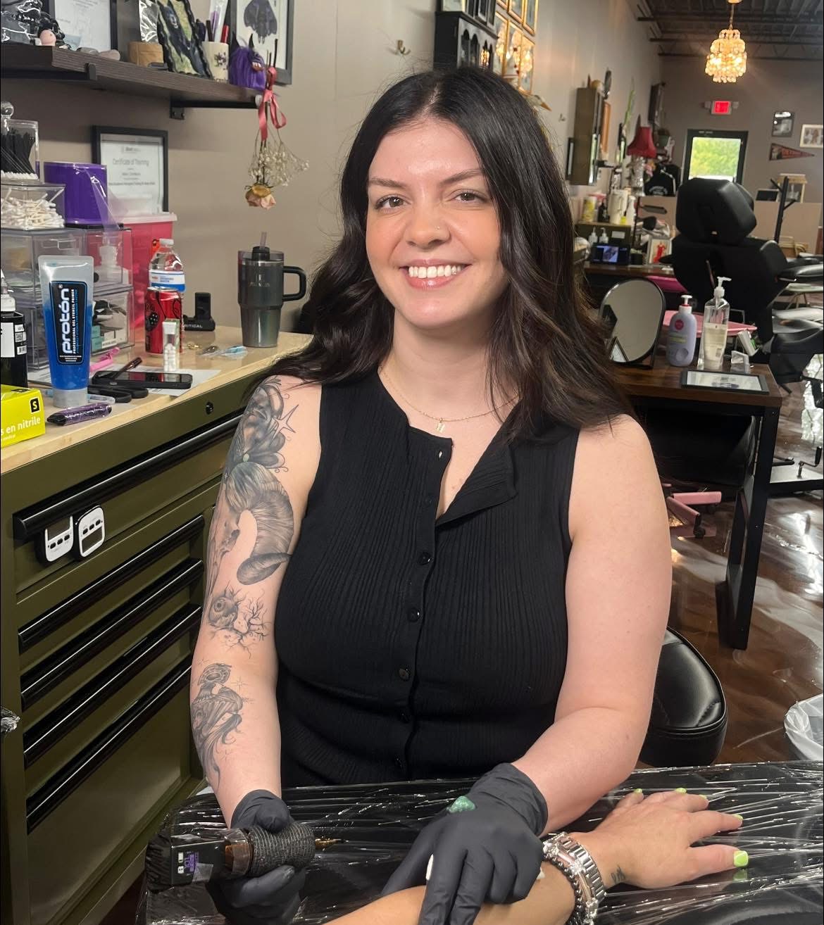 Woman with dark hair and tattoos smiling, sitting at a tattoo studio, wearing black sleeveless top and gloves.