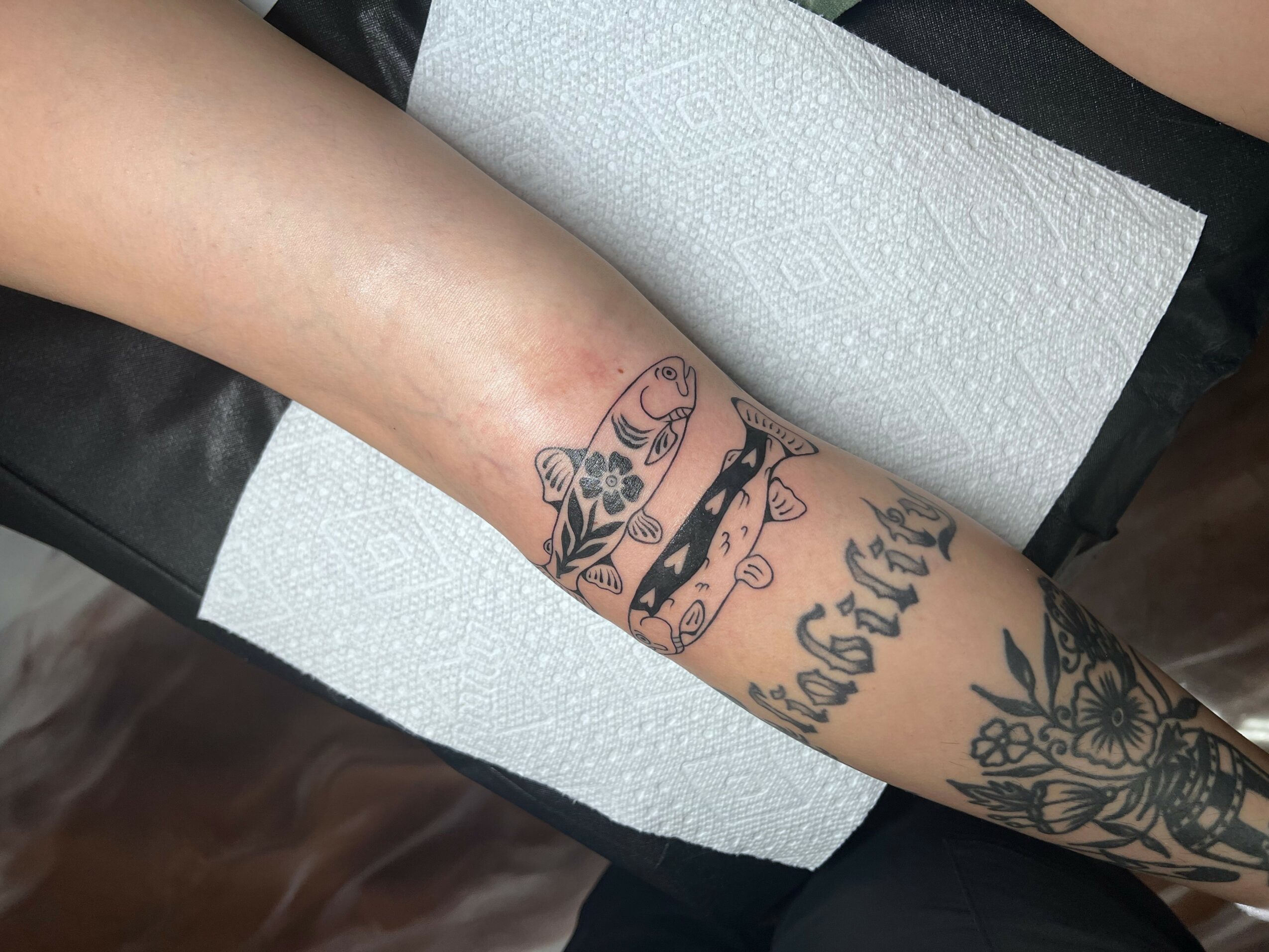 Tattooed arm with various designs, including a cat, flowers, and text, resting on a black surface with paper towels nearby.