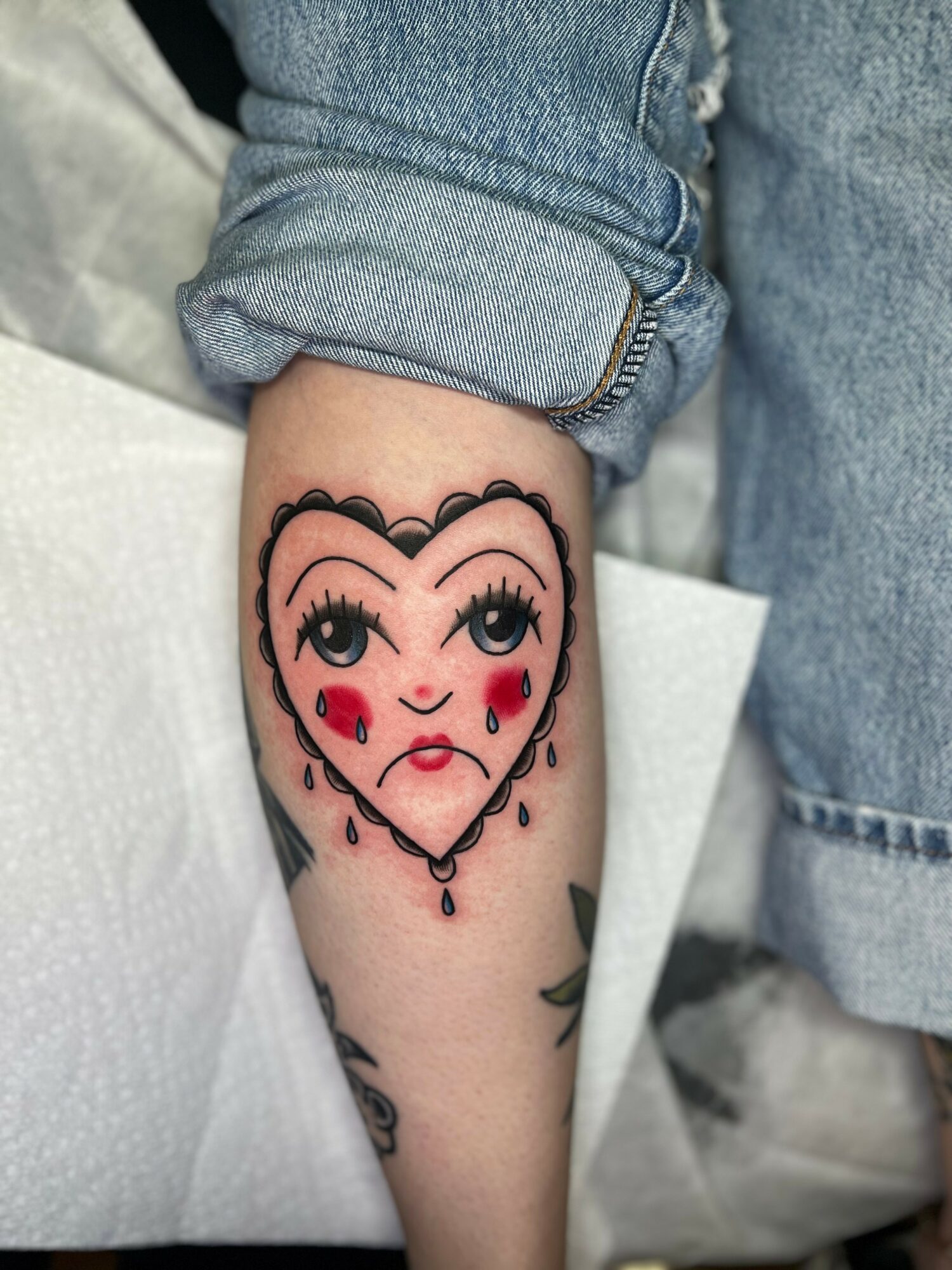 Tattoo of a sad face with tears, on a person's forearm, with rolled-up denim jeans visible.