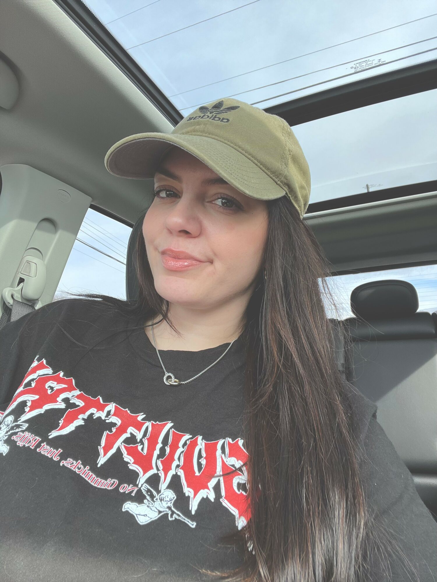 Young woman with long dark hair wearing a beige cap and black graphic T-shirt, sitting inside a vehicle with a sunroof.
