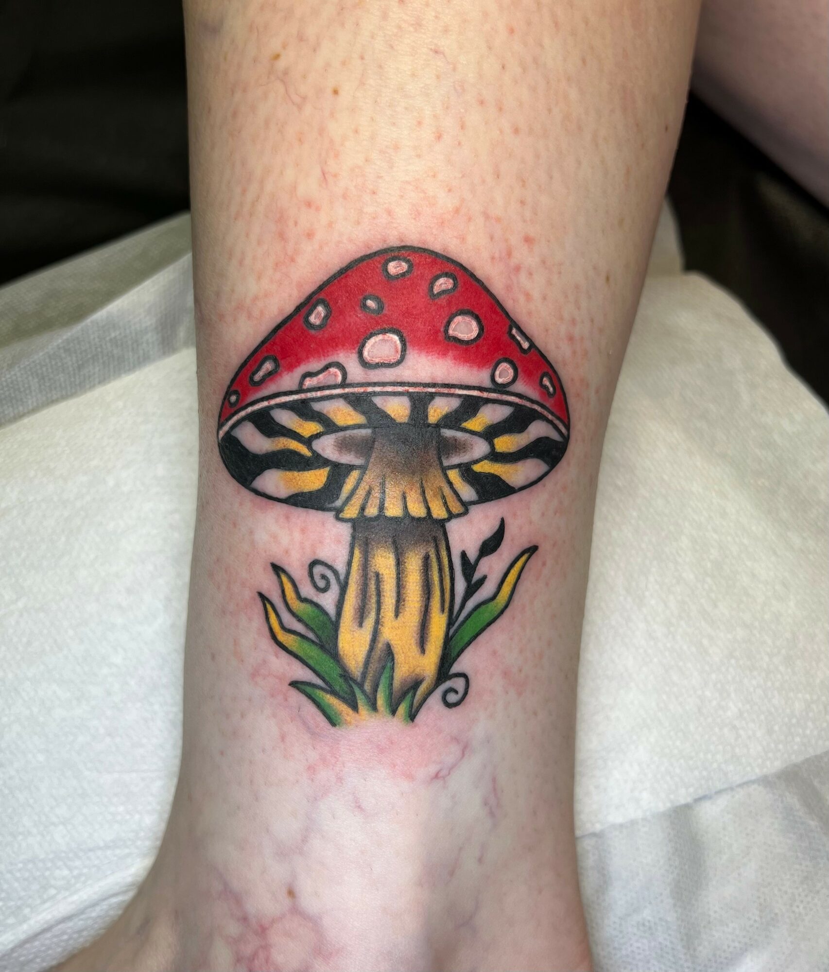Colorful mushroom tattoo with red cap, white spots, yellow stem, and green grass at the base, on skin.