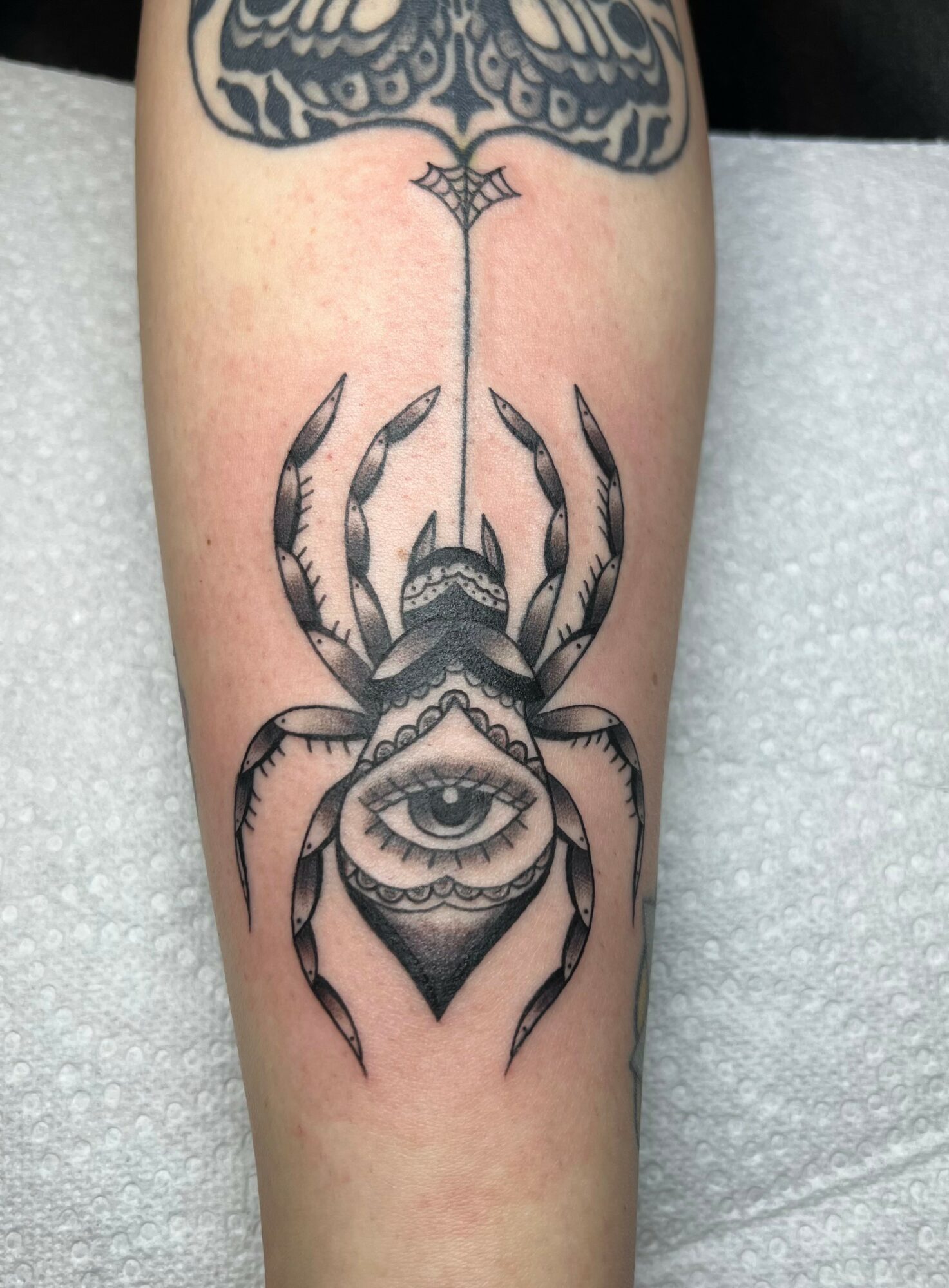 Tattoo of a crab with an eye inside a diamond shape on its body, featuring a vertical line with a web-like design above.