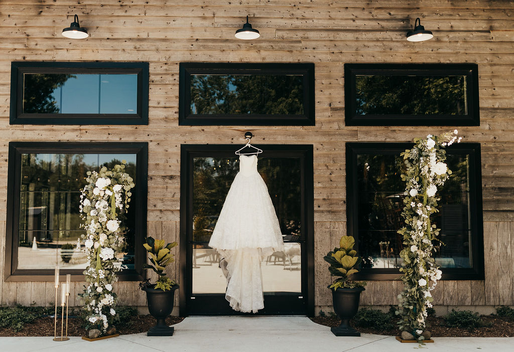 Wedding dress hanging in front of a building with three windows, flanked by floral arrangements and potted plants.