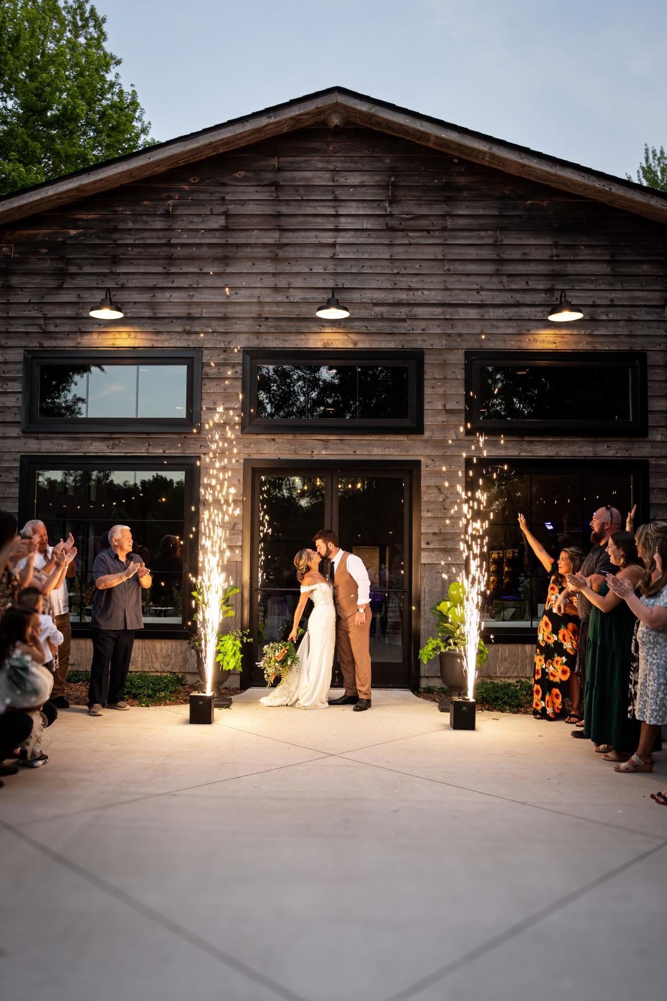 Couple kissing outdoors at wedding reception, surrounded by guests, with sparklers and a rustic building in the background.