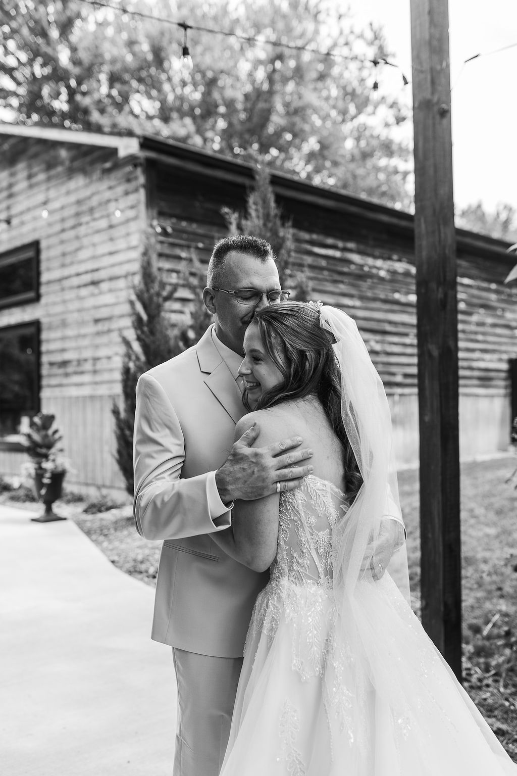 A bride and groom embrace outdoors, with the groom in a suit and the bride in a wedding dress, smiling.