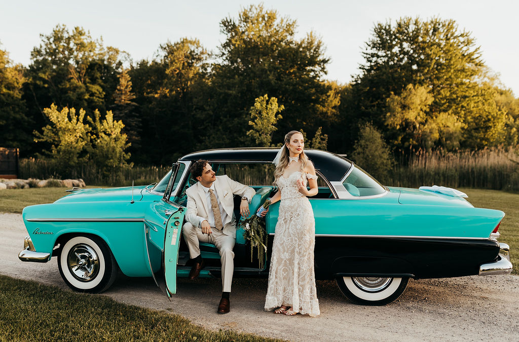 A man and woman in formal attire beside a vintage turquoise car outdoors with trees in the background.
