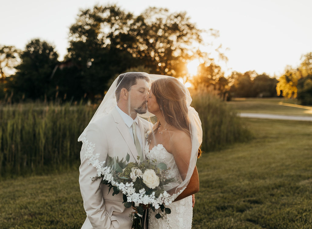A bride and groom stand close together outdoors at sunset, holding a bouquet of flowers, with trees and a grassy field in the background.