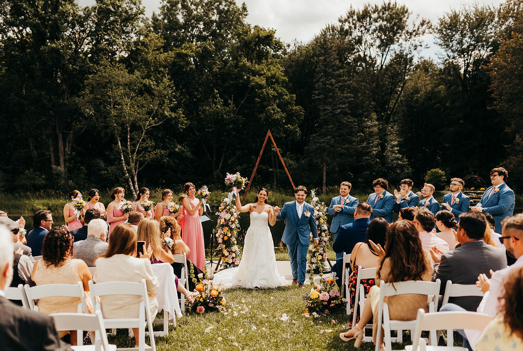 Outdoor wedding ceremony with bride and groom under an arch, surrounded by guests seated on white chairs, trees in background.