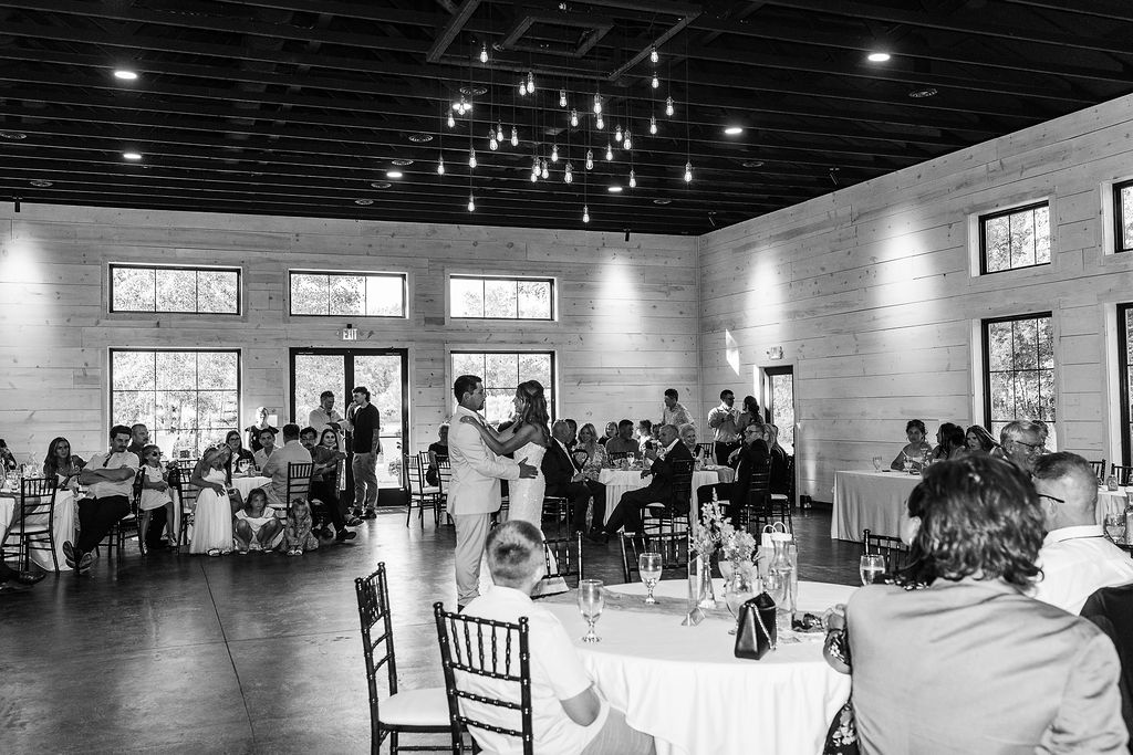 People gathered in a spacious room with large windows, sitting at round tables and standing, with a person dancing in the center.
