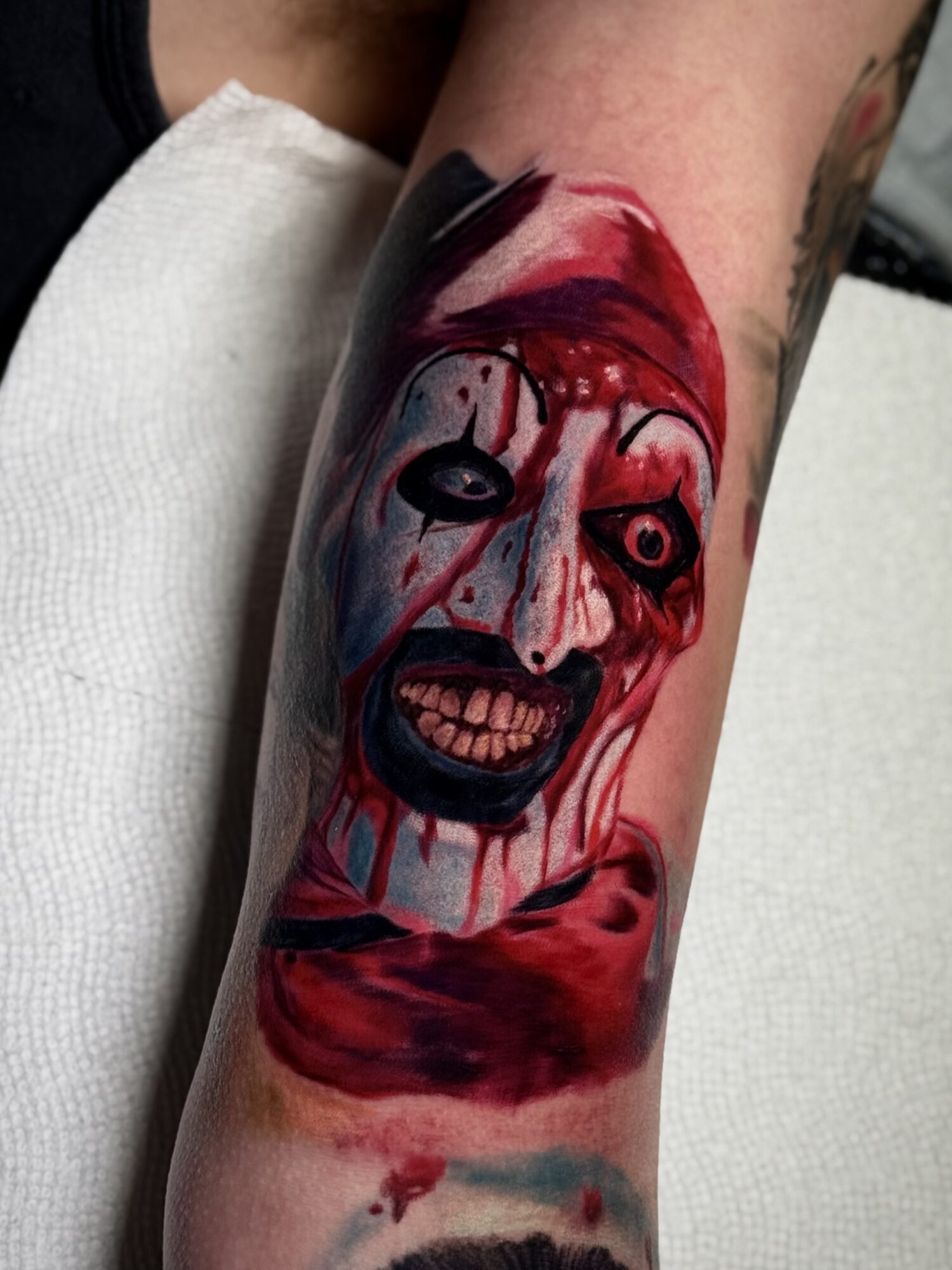 Tattoo of a scary clown face with blood and a sinister expression on a person's arm.