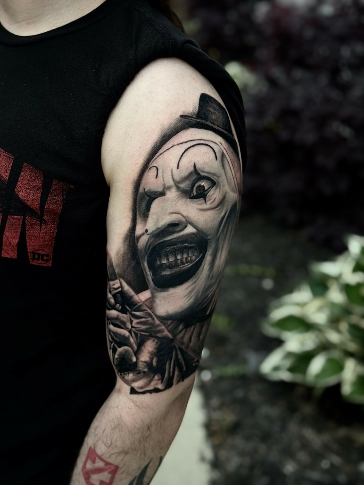 Tattoo of a clown face with exaggerated features on person's upper arm, with dark background and greenery nearby.