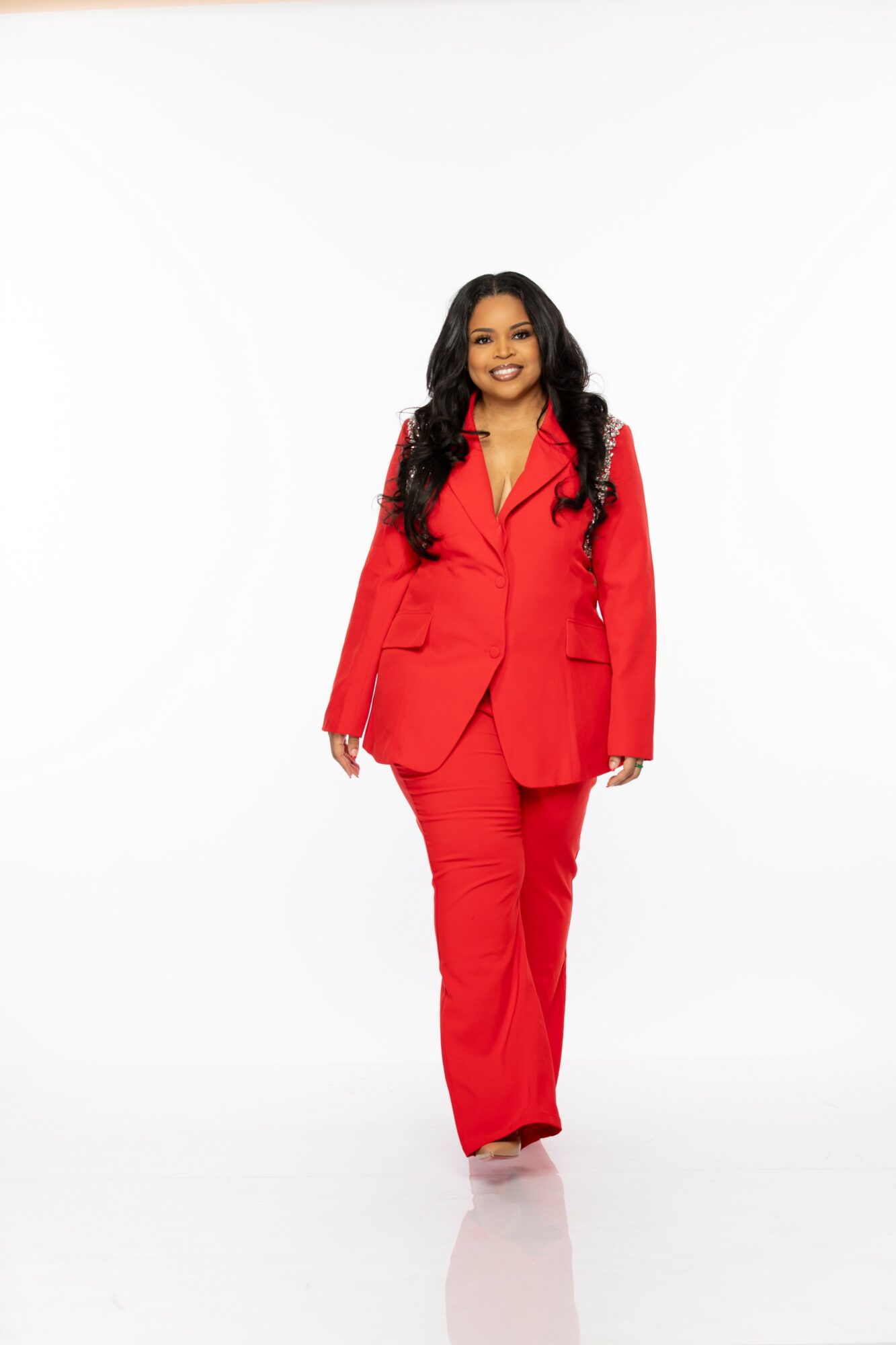 Woman in a red suit walking towards the camera, smiling, with a white background.