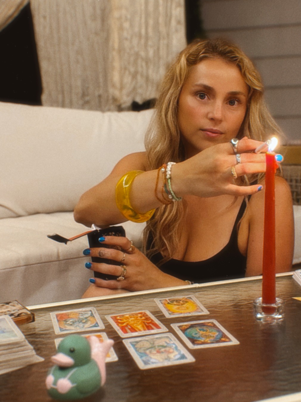 Woman with long blonde hair holding a lit candle, sitting at a table with tarot cards, in a cozy room.